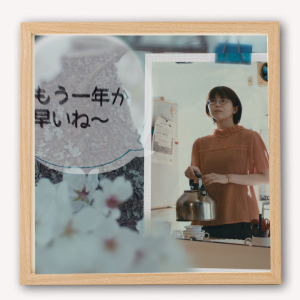 カフェのレジ横の鏡越しに女性が映り、一緒に文字入り泡タイプのサインボードと花の飾りも見える