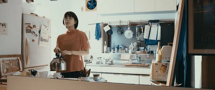 キッチンにいる女性がコーヒーポットを持っている。背景には冷蔵庫や調理器具、飾り棚が見える。
