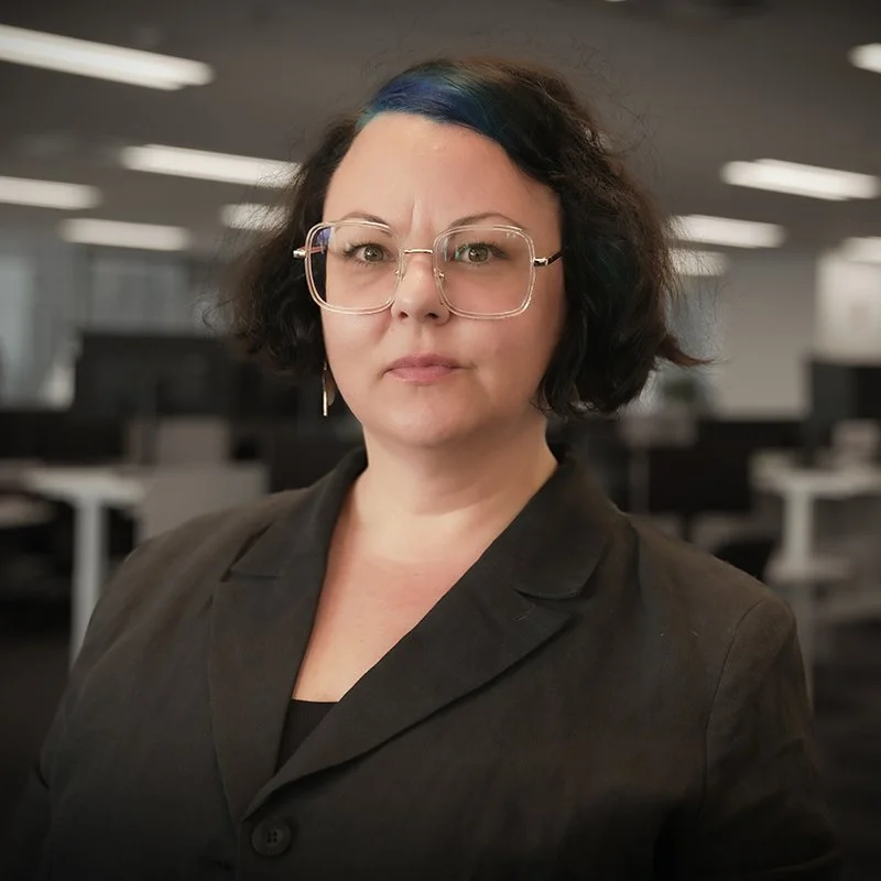 Bronya has light skin, short brown hair and brown eyes. She is wearing a black shirt and there is a corporate office in the background.