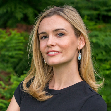 Rebecca Lake has light skin, long blonde hair and blue eyes. She is wearing a dark blue shirt and there are trees in the background.