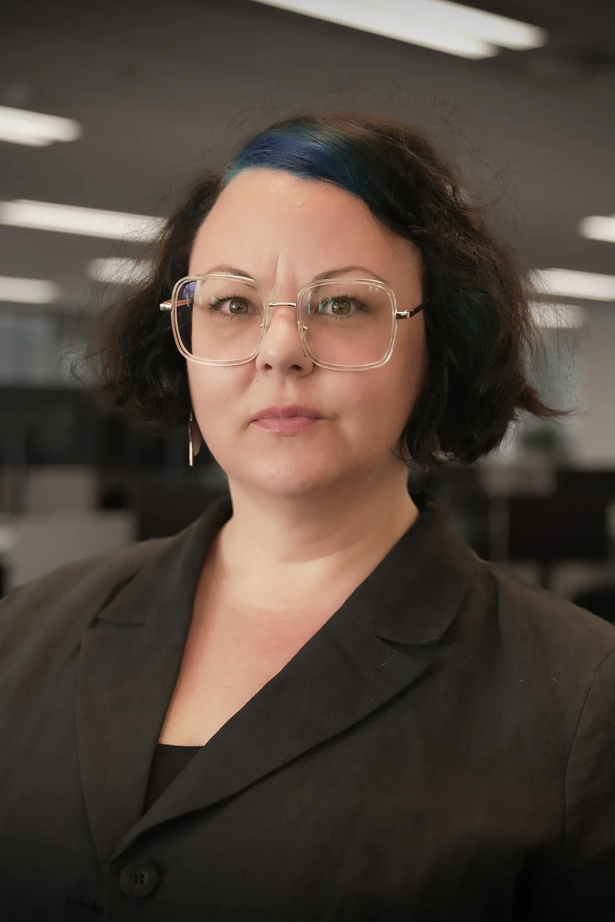 A woman with shoulder-length dark hair and light streaks, wearing glasses with large clear frames, and a black blazer, standing in an office environment.
