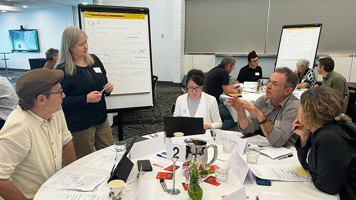 People sitting around a conference table engaged in discussion, with a woman standing and writing on a whiteboard in a classroom or conference room setting.