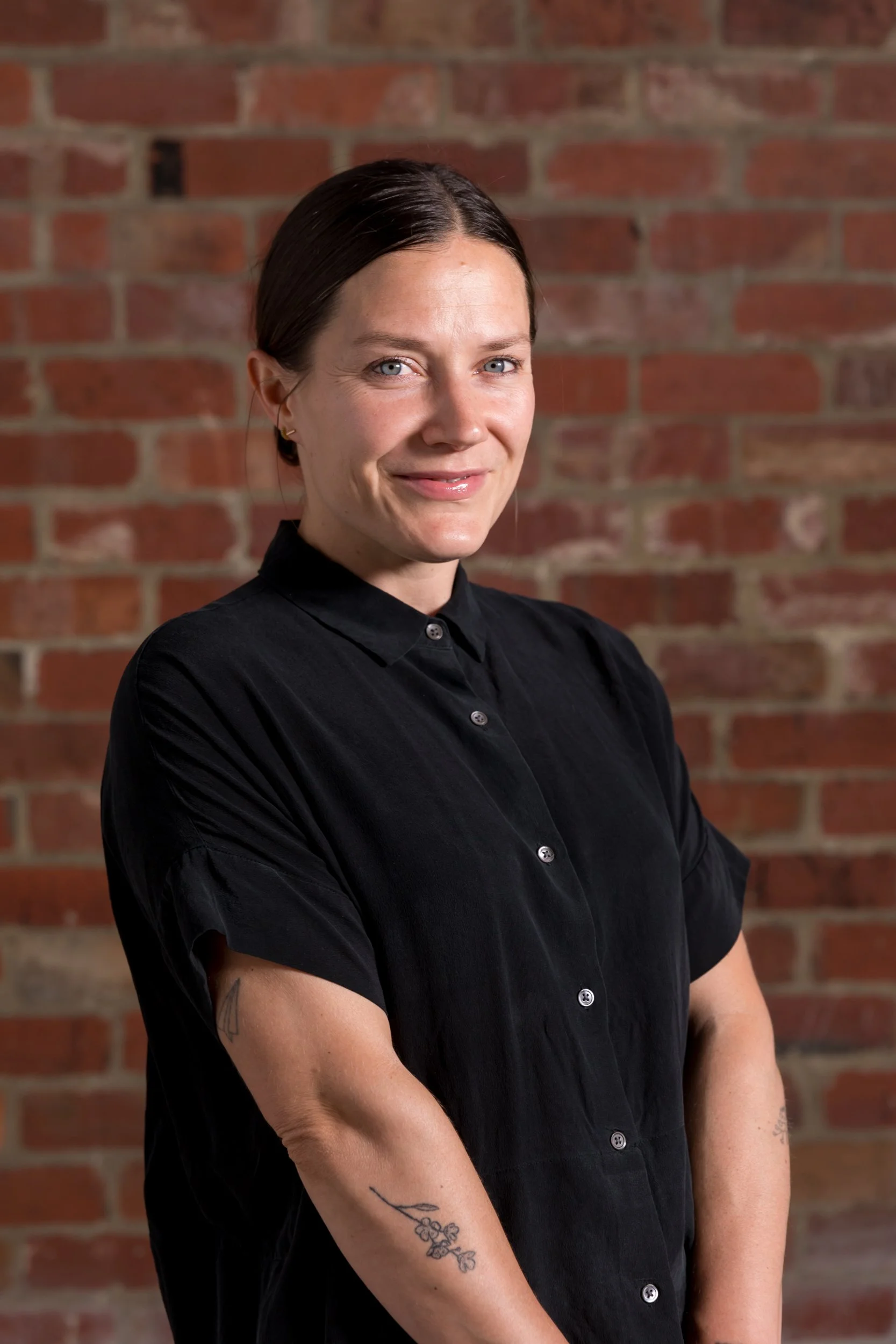 Martine Holberton, Founder. Martine has dark brown hair, blue eyes and light skin. She is smiling and wearing a black button-up shirt while standing in front of a red brick wall.