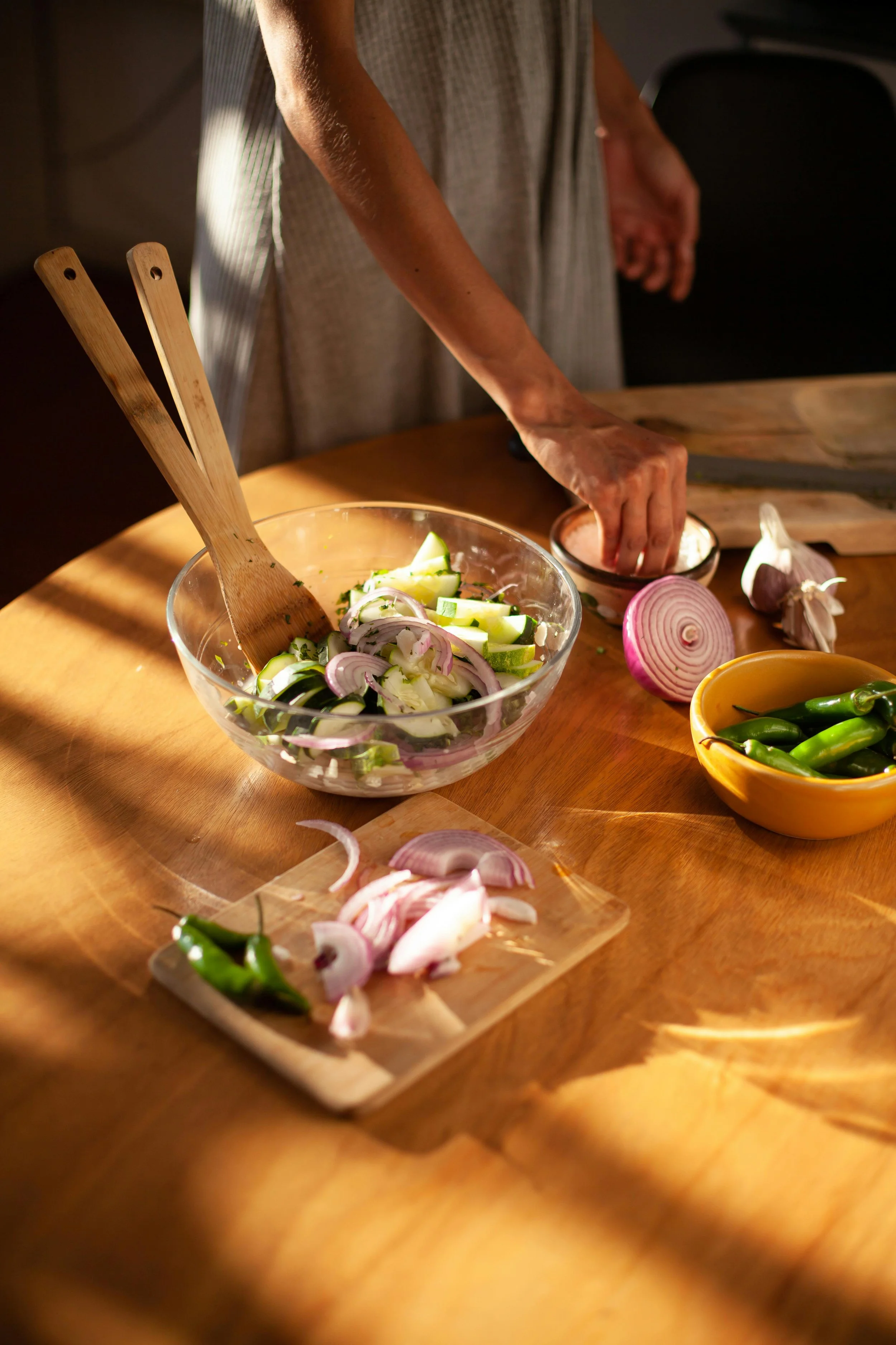Person chopping onions and preparing vegetables on a wooden cutting board, with a bowl of sliced vegetables and a bowl of chili peppers nearby.