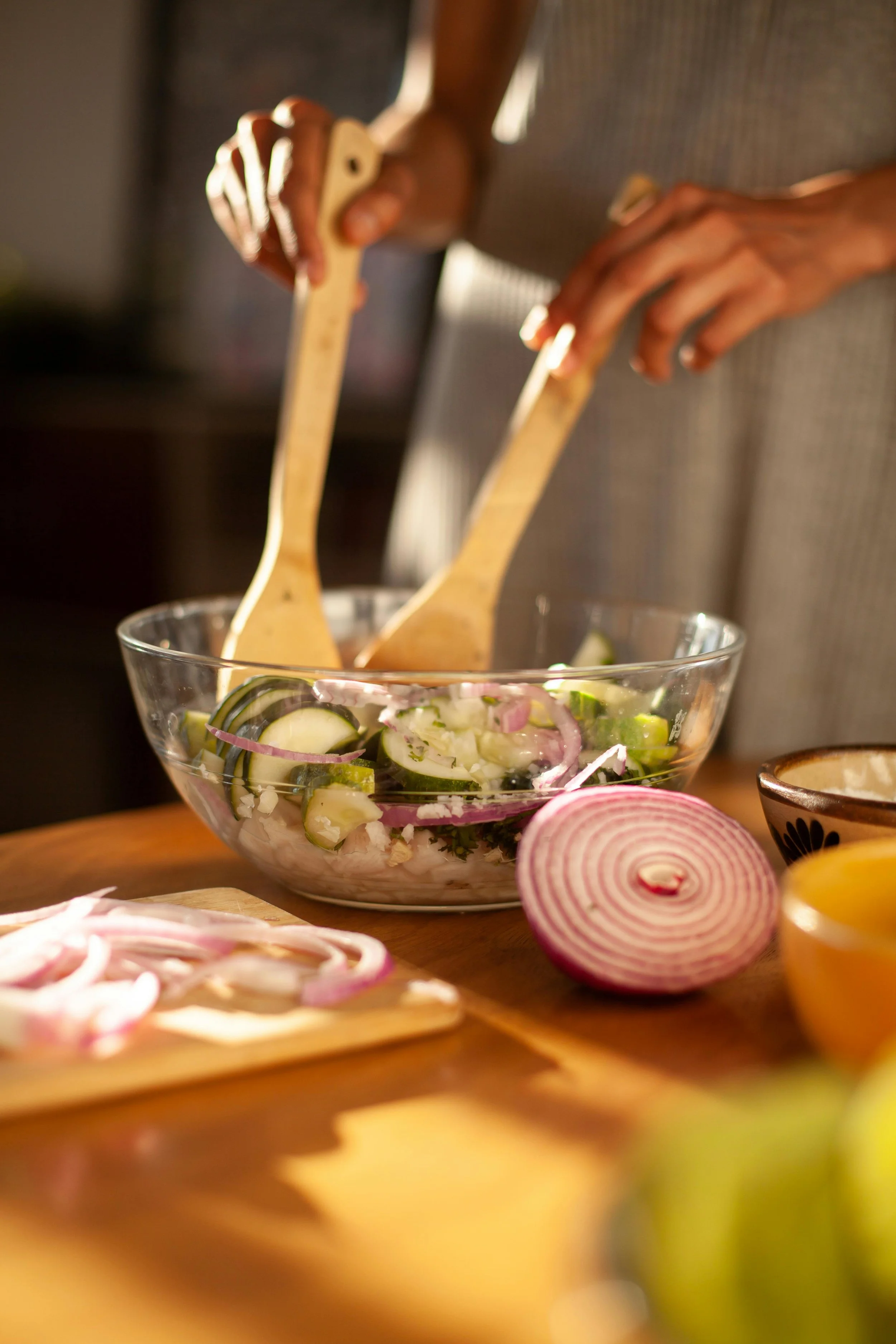 Close-up of a hand holding wooden salad servers above a glass bowl containing a cucumber, onion, and parsley salad, with sliced onions and other vegetables on a wooden cutting board nearby.