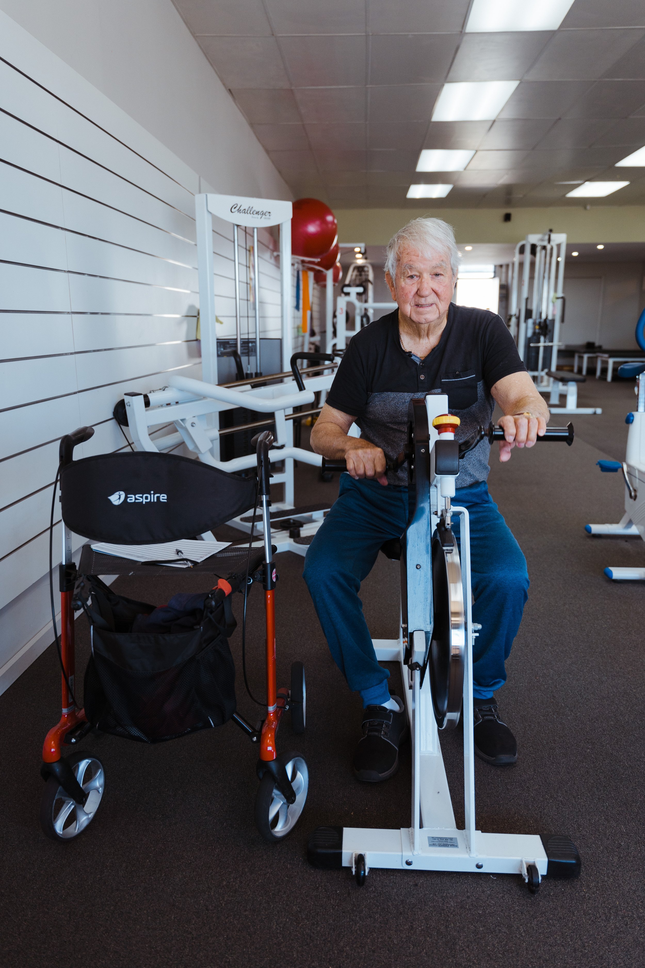 An elderly man with gray hair exercising on a stationary bike in a gym. A wheelchair is nearby, and gym equipment is in the background.