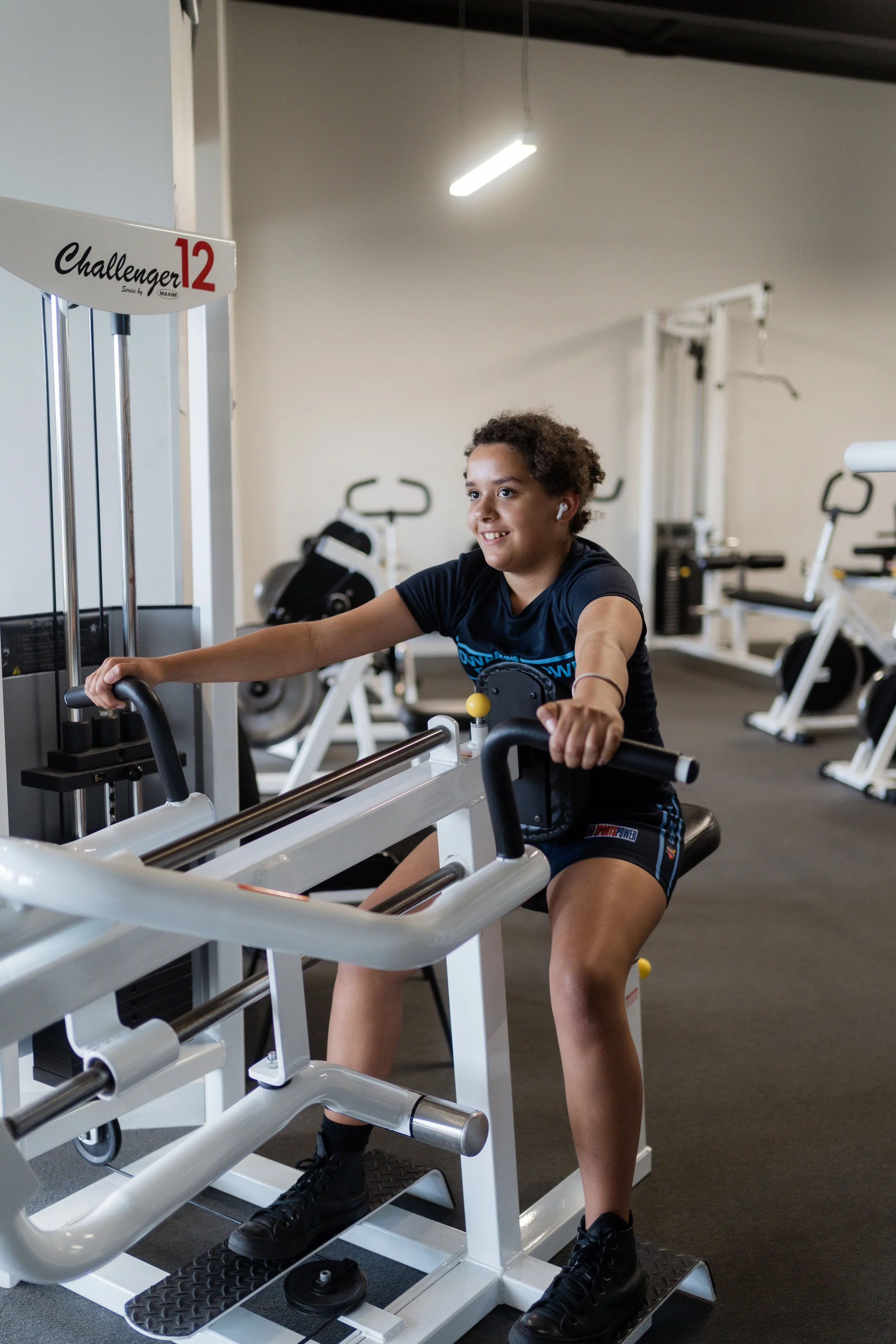 A young girl with curly hair and AirPods exercising on a leg extension machine at a gym.