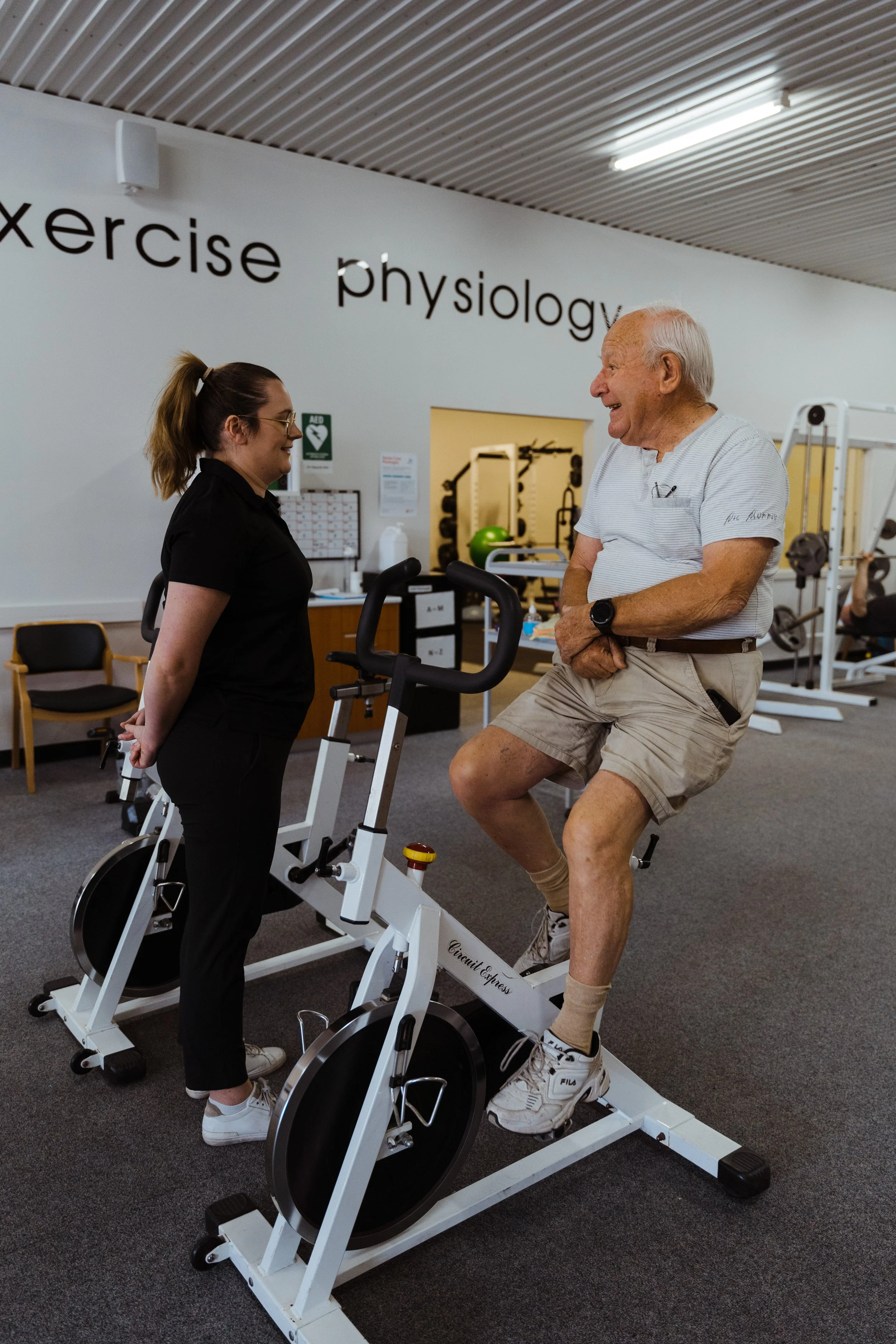 An elderly man with a white shirt and shorts exercising on a stationary bicycle in a gym, talking with a woman in black who is standing nearby. The gym has medical or exercise equipment and a sign on the wall that reads 'exercise physiology.'