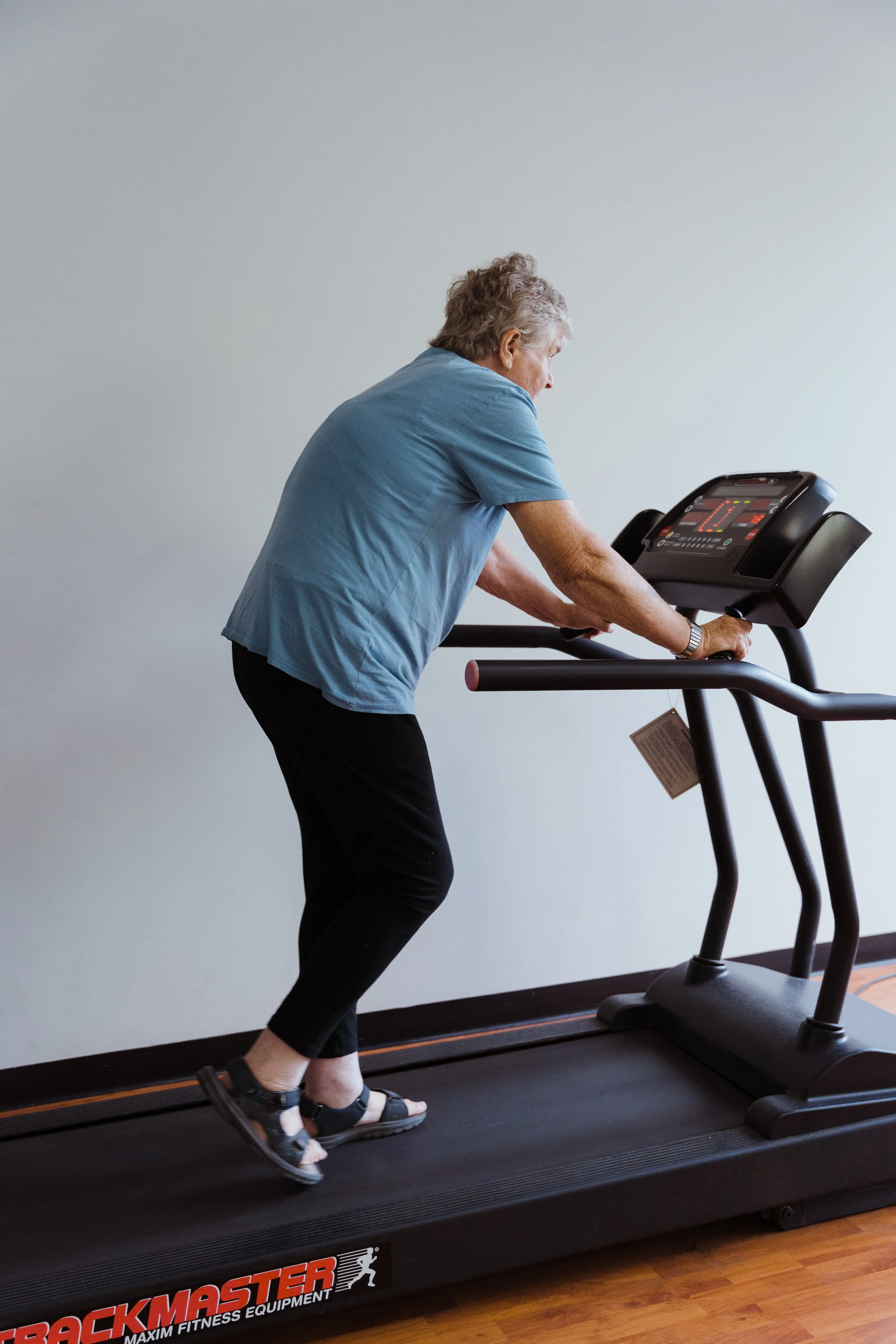 An elderly woman with gray hair, wearing a blue shirt and black pants, walking on a treadmill in a gym.