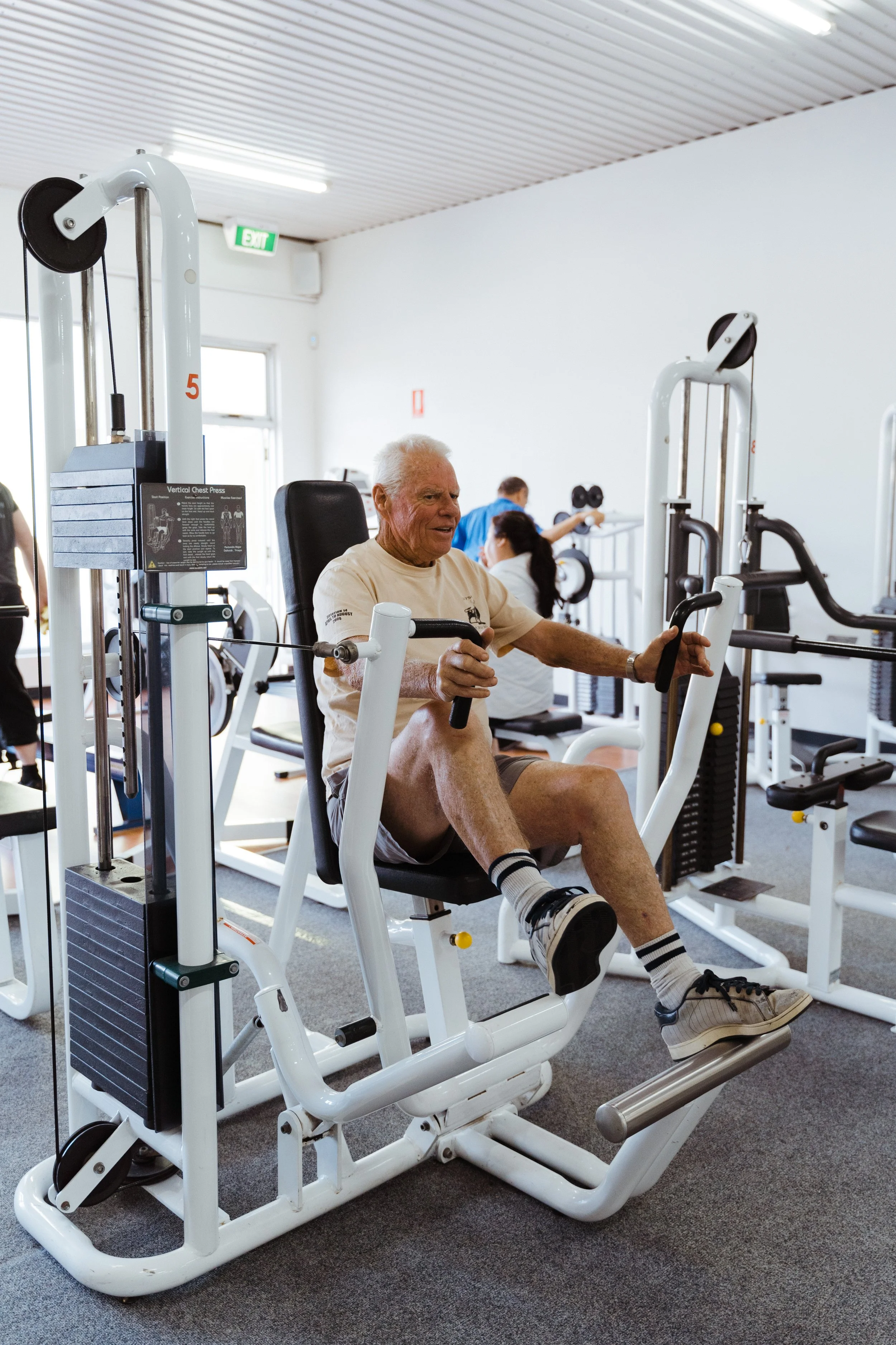 An elderly man exercising on a leg press machine in a gym, with other people working out in the background. The gym has white walls, a gray carpet, and a ceiling with fluorescent lighting.