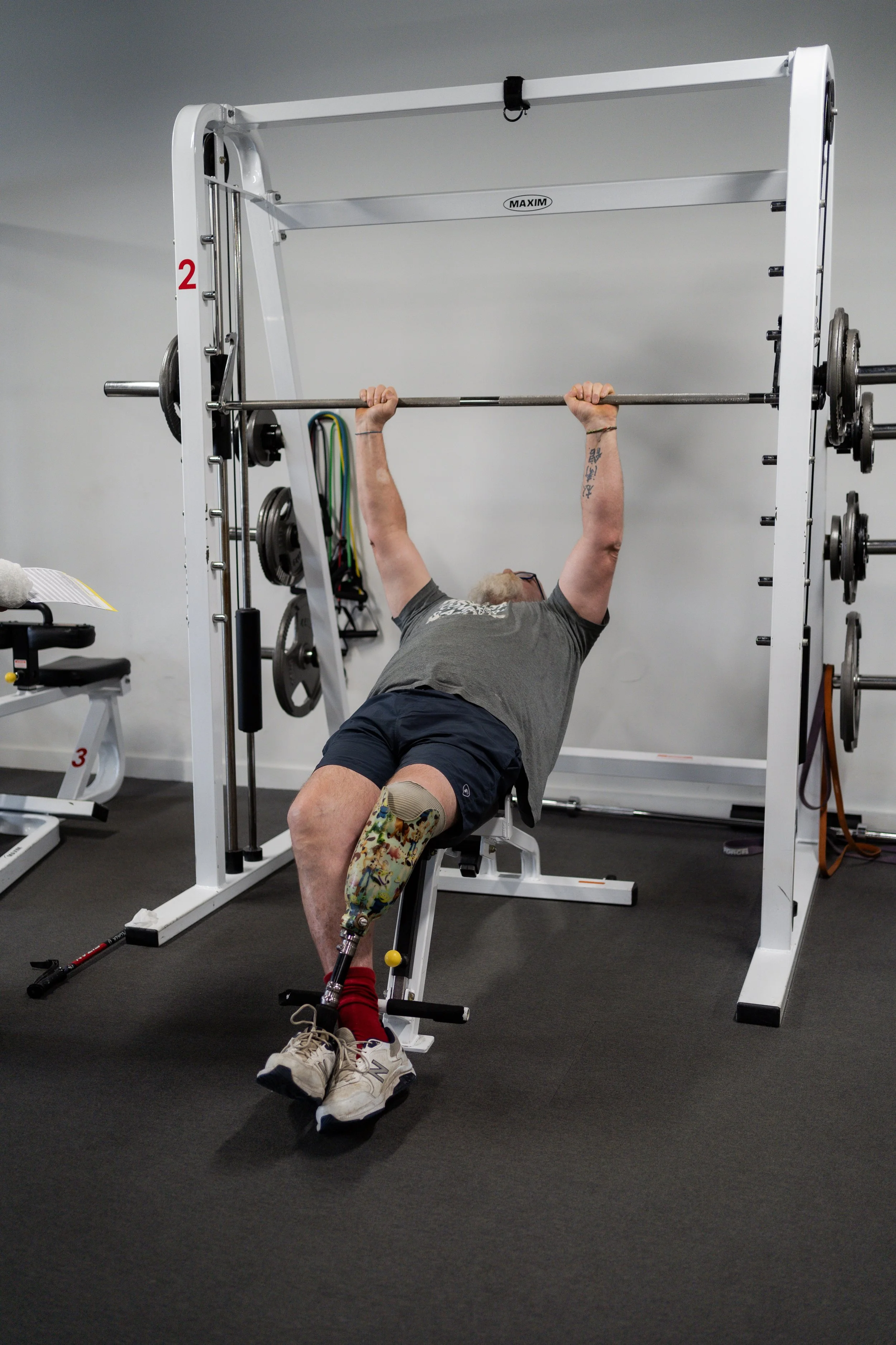 A man with a prosthetic leg lifting a barbell in a gym, lying on a weight bench. The gym has weight plates, resistance bands, and workout equipment around him.