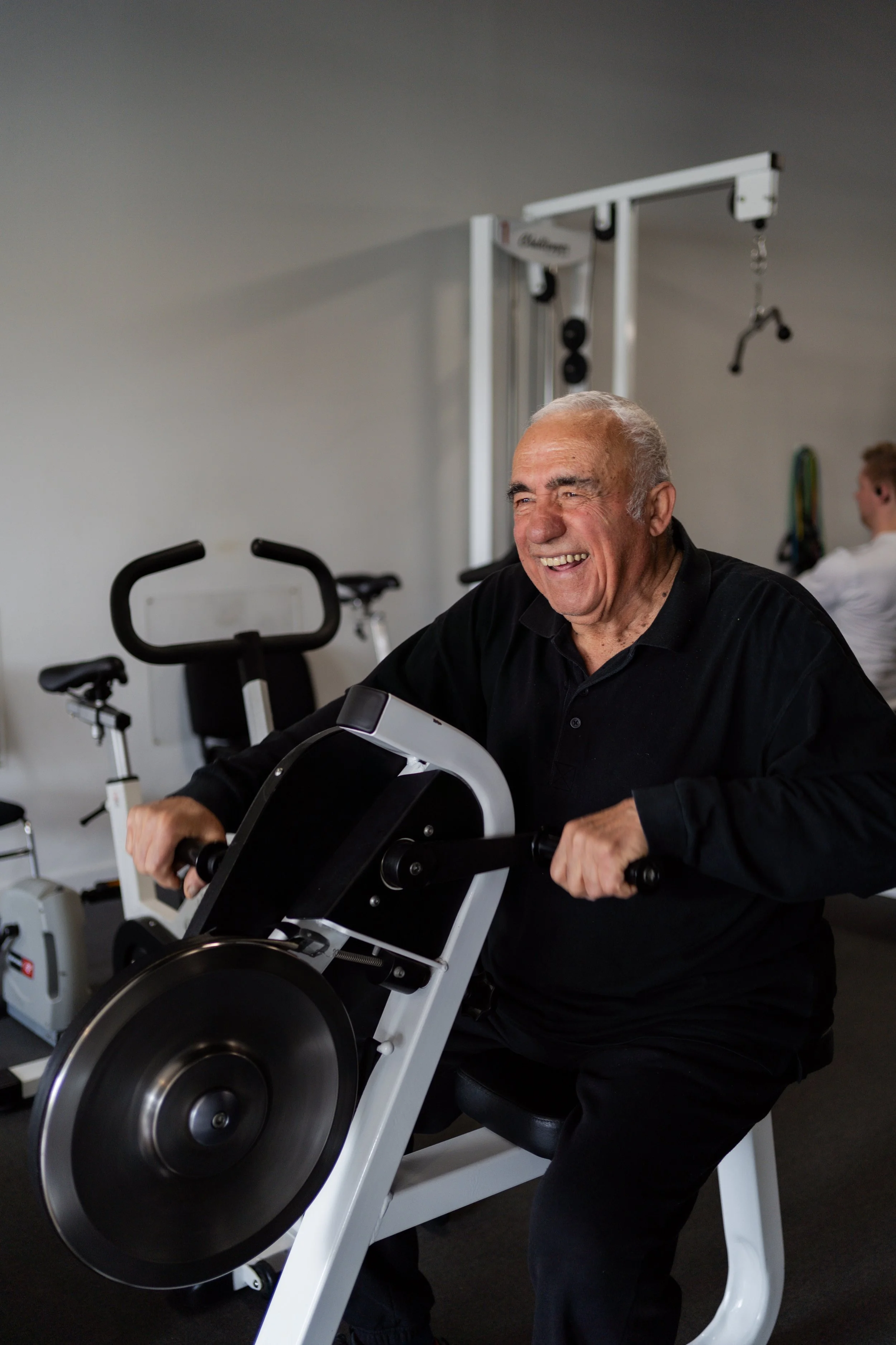 An elderly man smiling and exercising on a wheelchair bike in a gym
