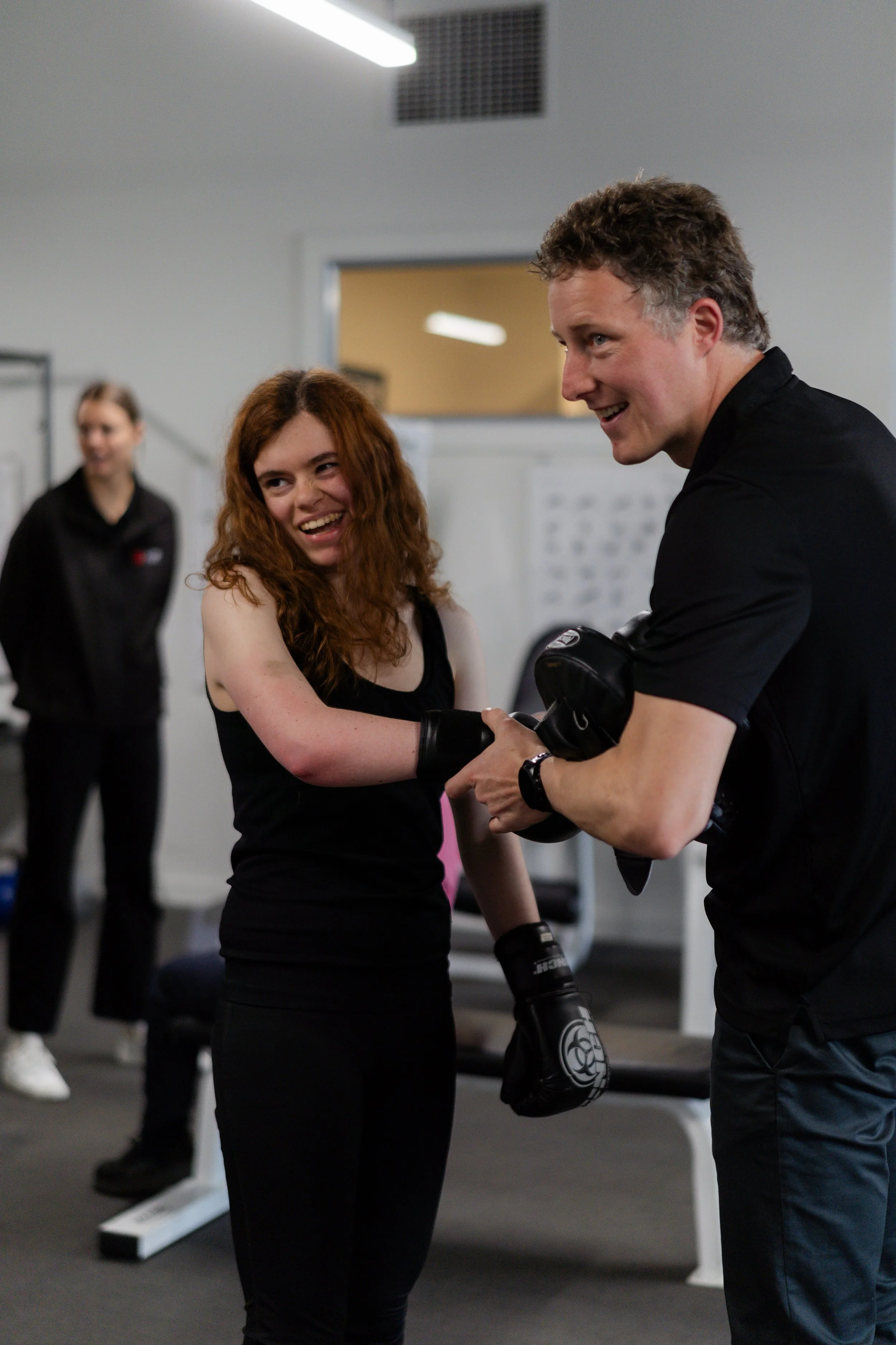 A young woman with long curly red hair wearing a black tank top and gloves, shaking hands with a man in a black shirt and boxing gloves in a gym, with a woman in the background watching.