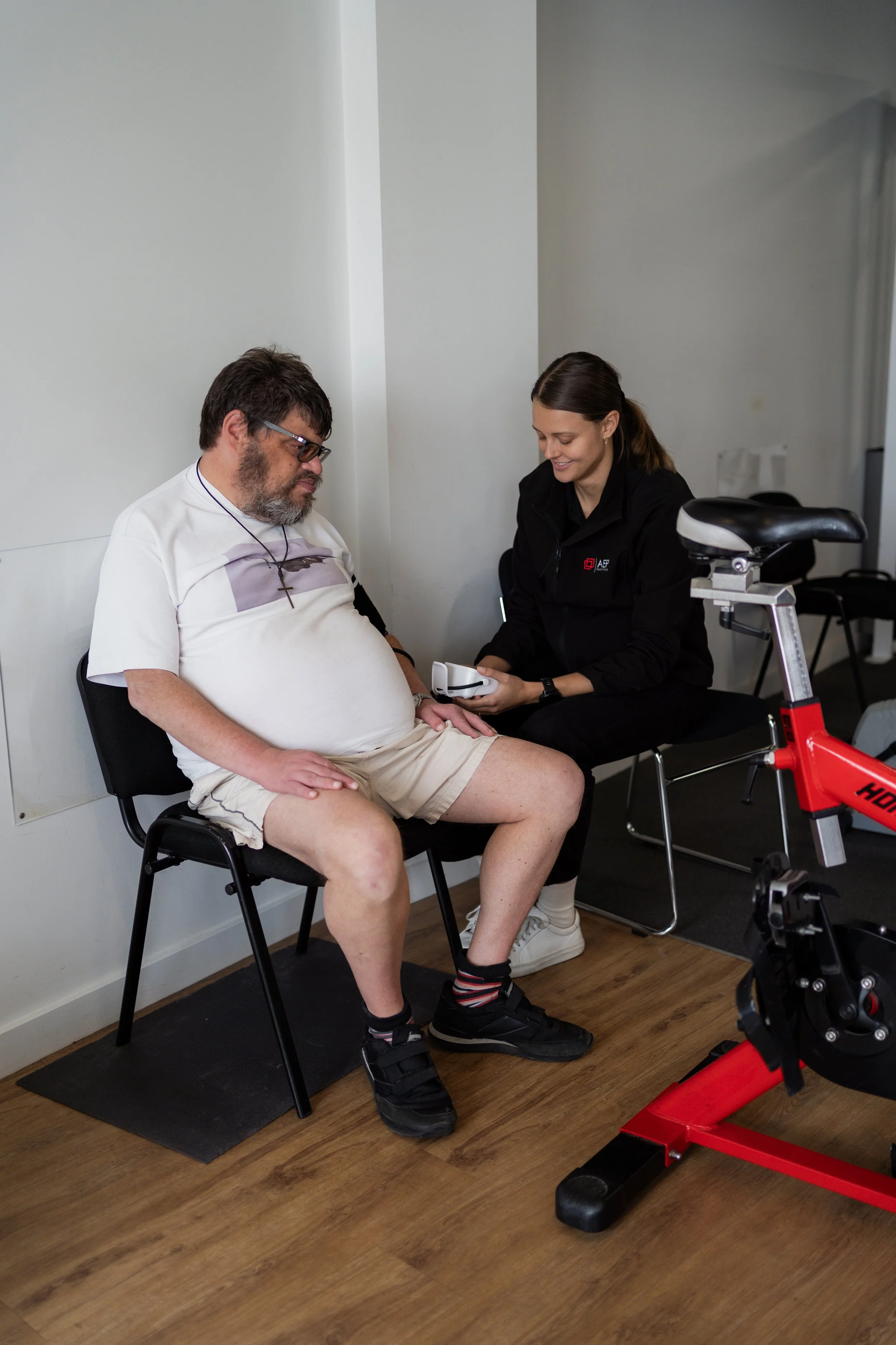 A man sitting in a chair wearing a white t-shirt, beige shorts, and black sneakers with socks, with a cross necklace around his neck. A woman dressed in black, smiling, is showing him something while holding a remote control, in a room with wooden flooring and a red stationary bike in the foreground.