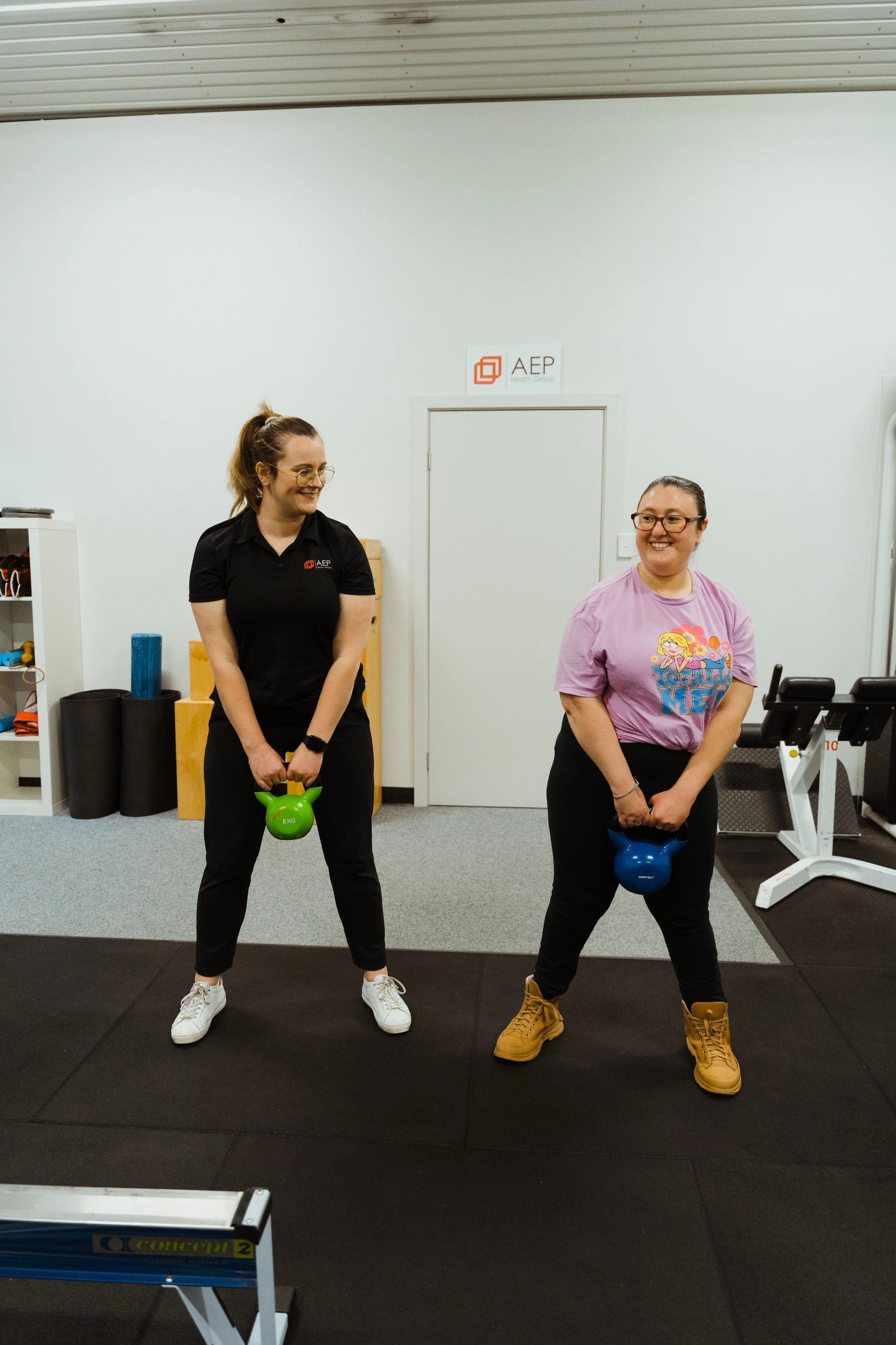 Two women standing in a gym, holding kettlebells, smiling and looking at each other.
