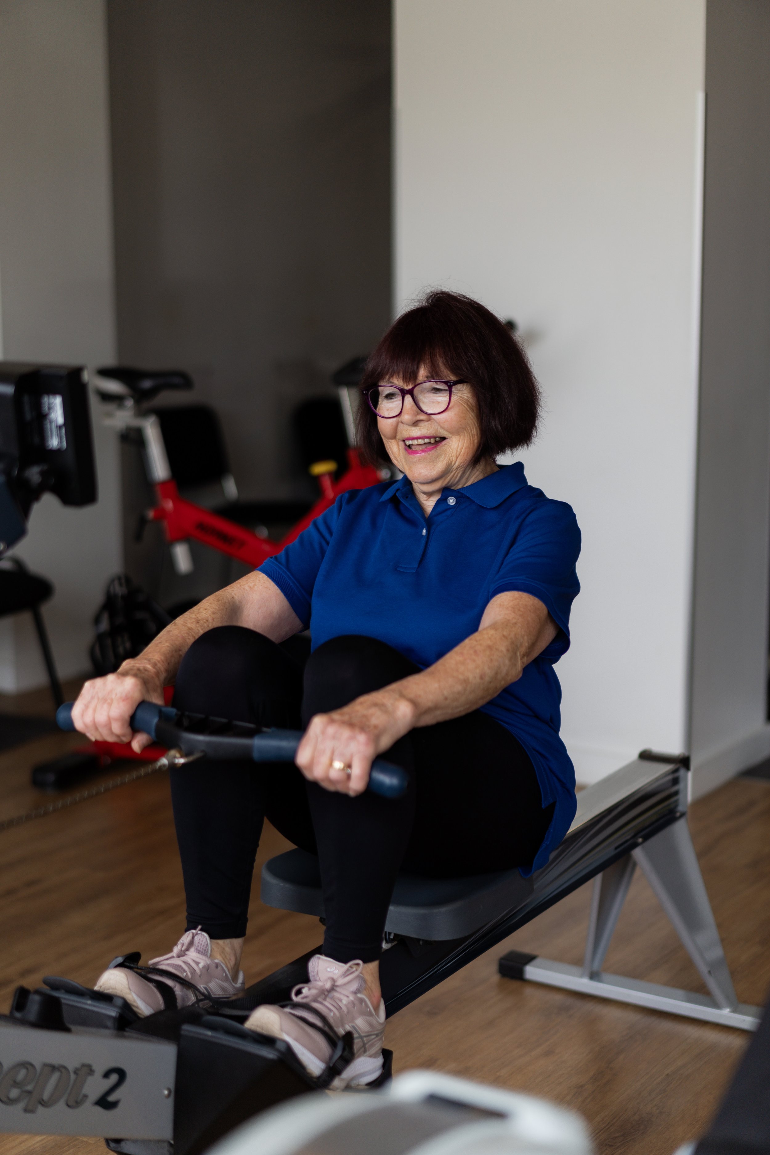 An elderly woman smiling while exercising on an elliptical machine in a gym, wearing a blue polo shirt, black pants, and pink sneakers.