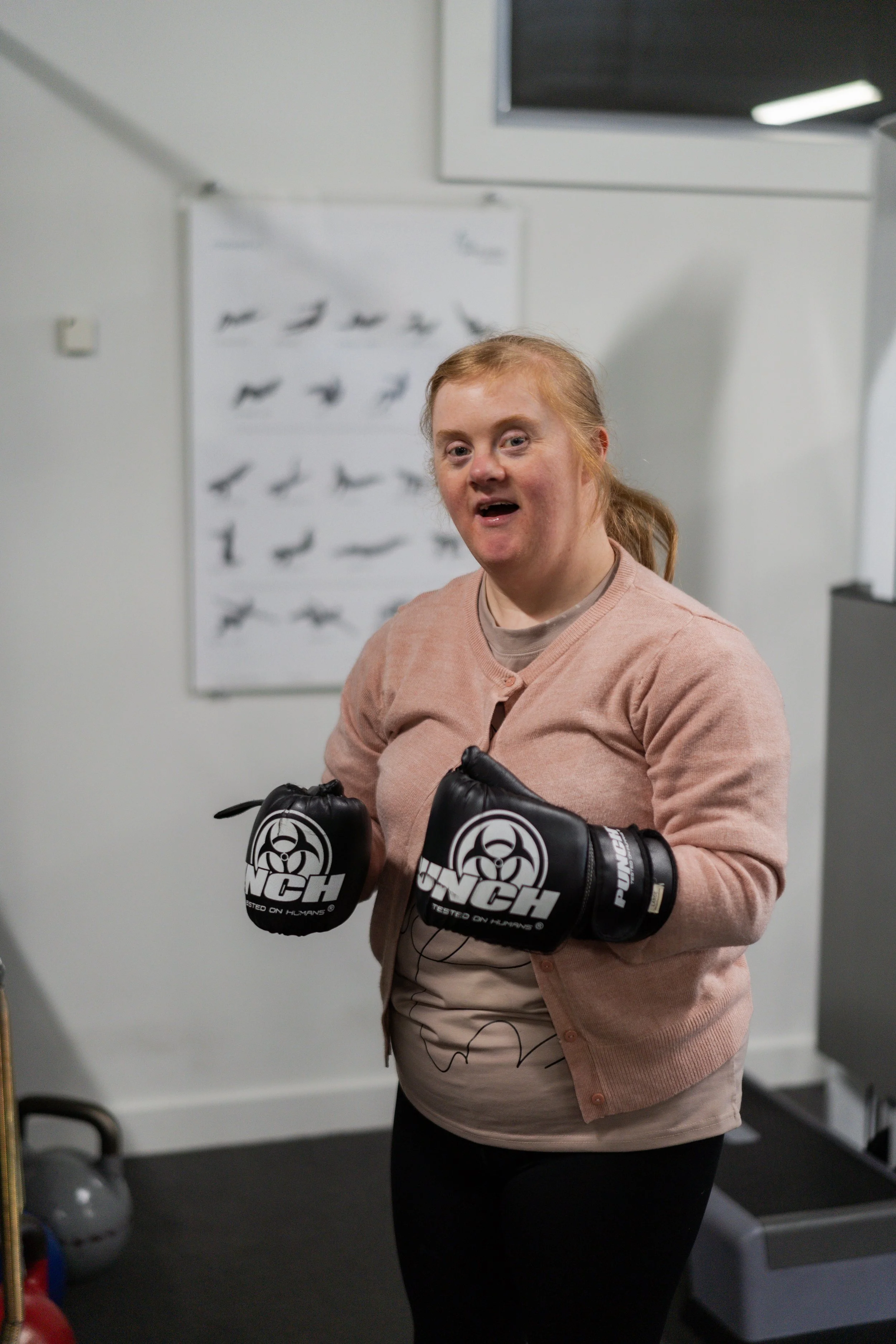 A woman with red hair in a ponytail, wearing a pink sweater and black pants, is practicing boxing with black boxing gloves in a gym. She is smiling and standing in front of a white wall with a poster of different boxing punches.