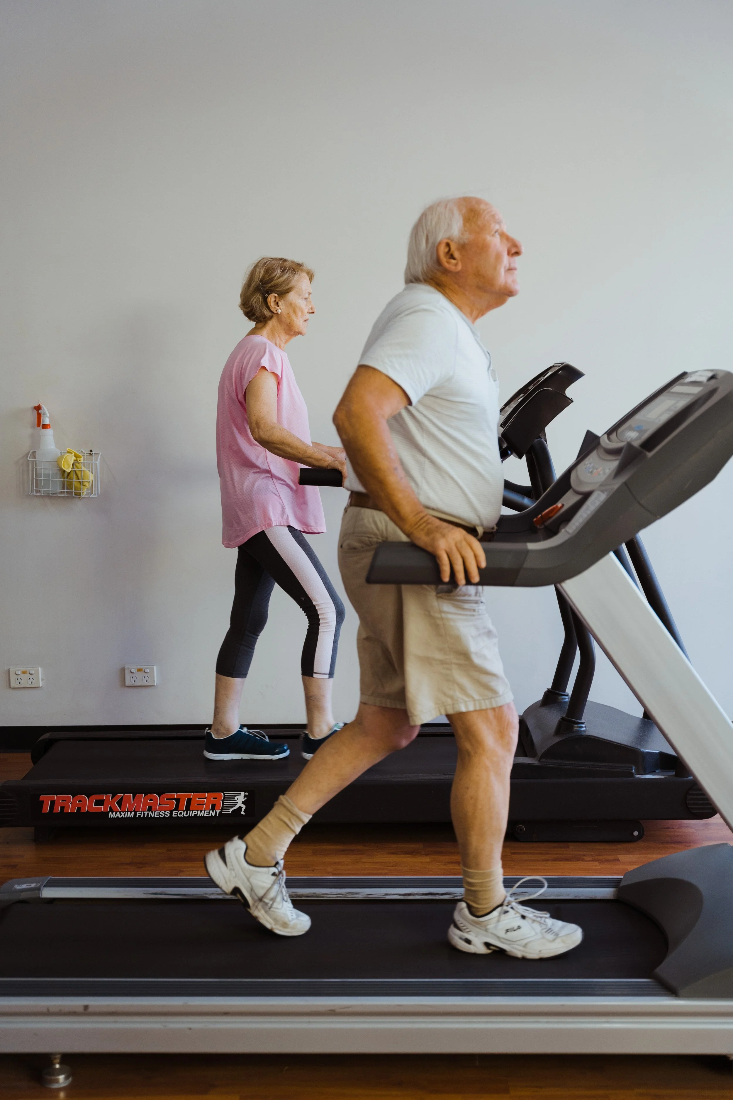Older man and woman walking or jogging on a treadmill in a gym, with minimalistic white wall background and cleaning supplies on the wall.