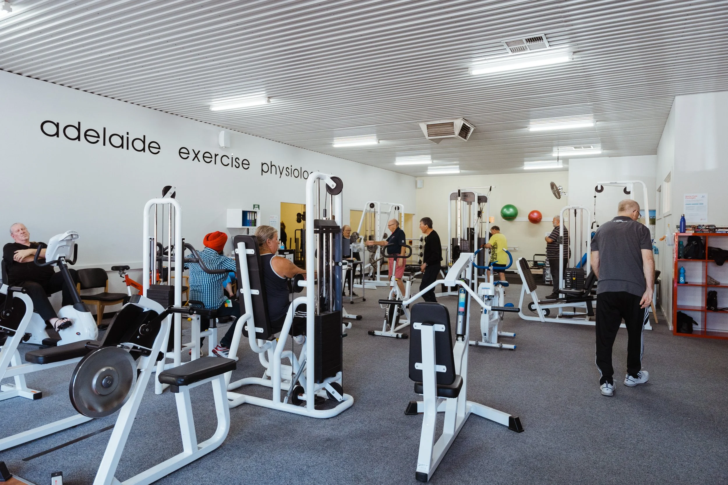 People exercising with cardio and strength training equipment in a gym, including ellipticals, stationary bikes, and weight machines, with a sign on the wall reading 'adelaide exercise physiolog'.