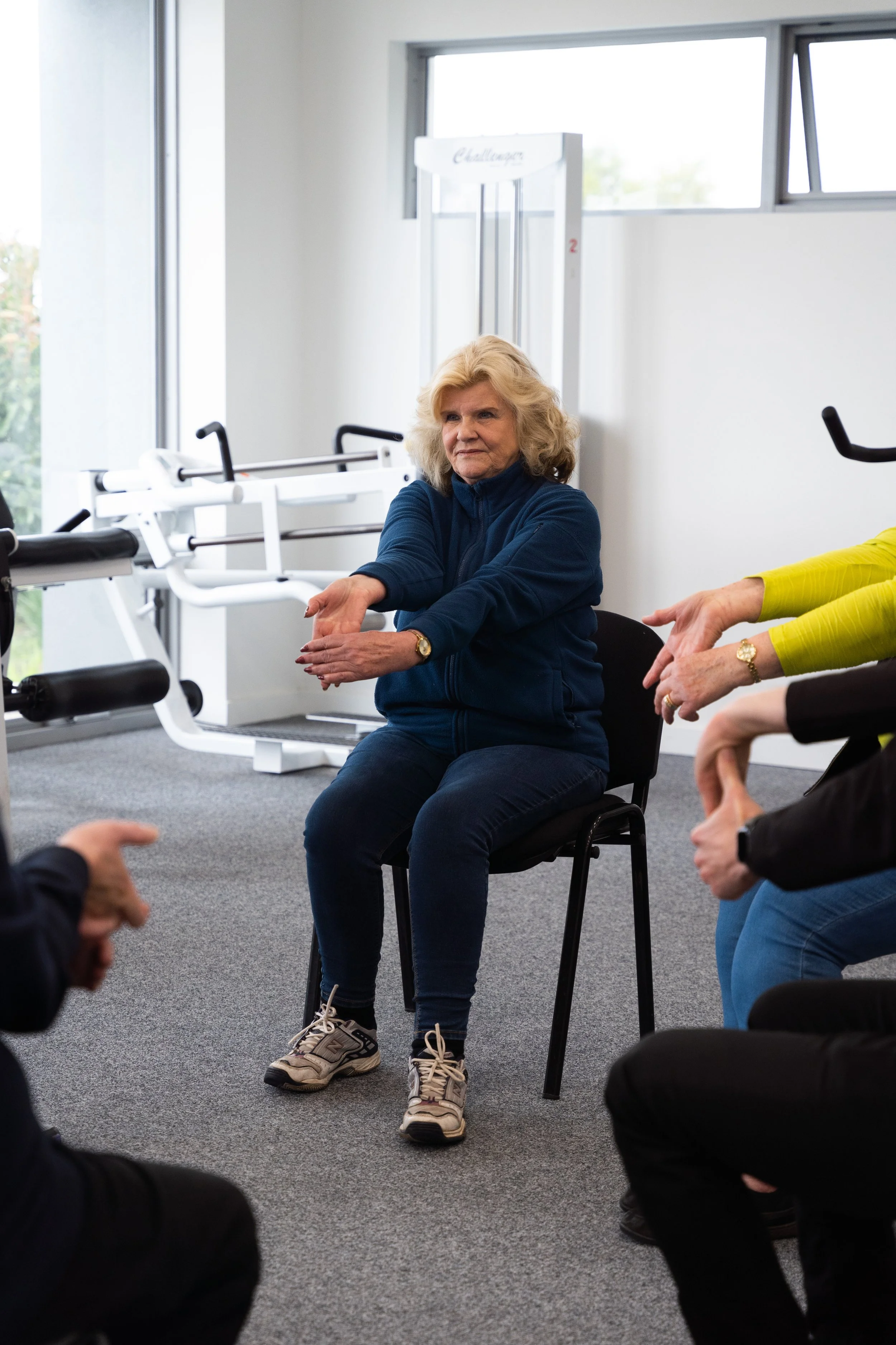 Older woman with blonde hair and a blue zip-up jacket, sitting on a chair and stretching her arms forward in a fitness or therapy session in a room with exercise equipment and large windows.