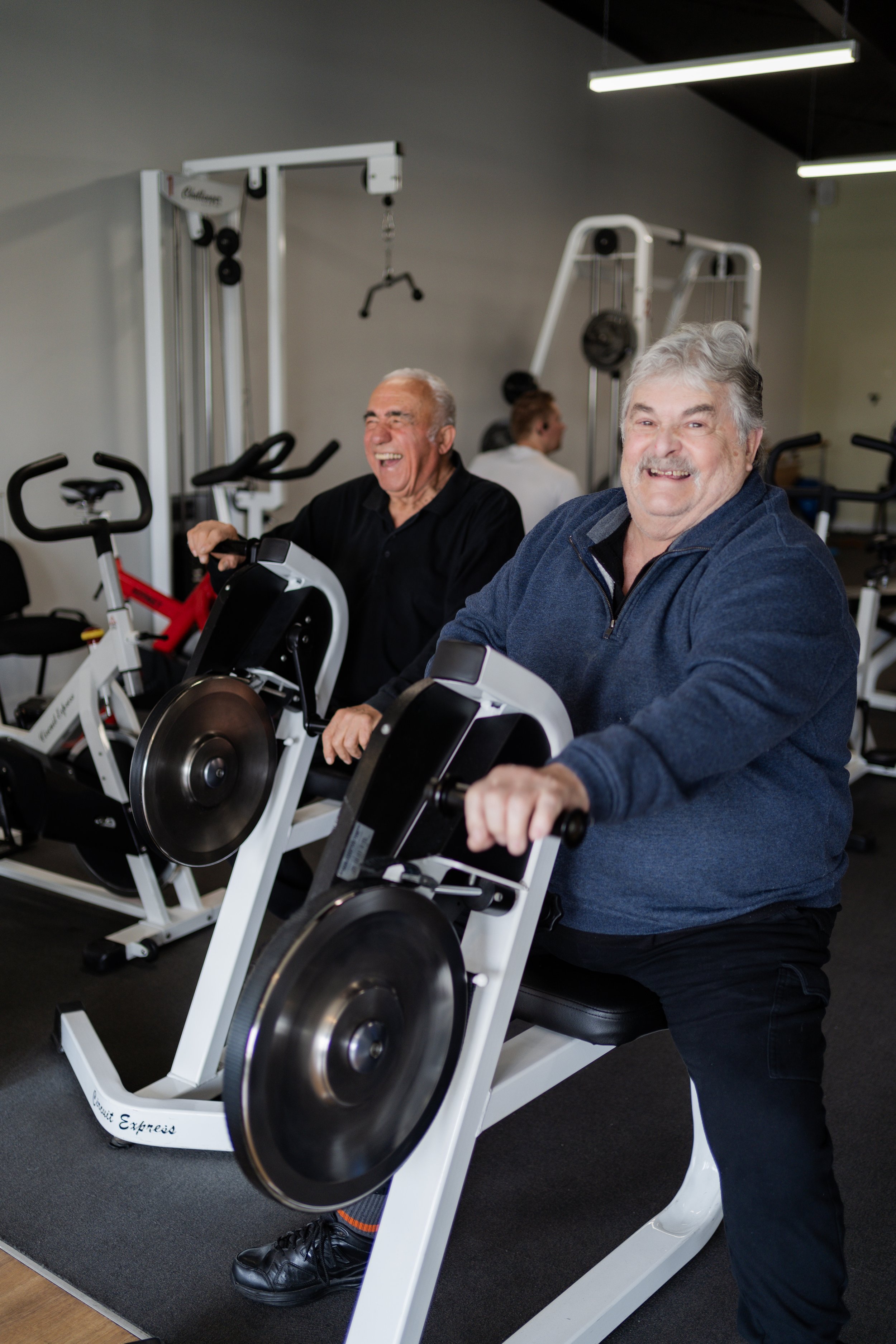Two elderly men exercising on stationary bikes in a gym, smiling and enjoying their workout.