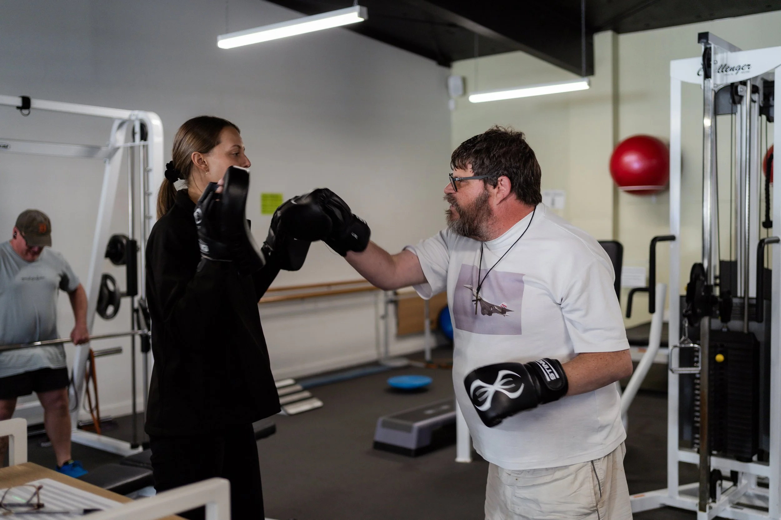 A man with glasses and a beard training a young woman in boxing at a gym. The woman wears boxing gloves and listens to the trainer. Other people are working out in the background.
