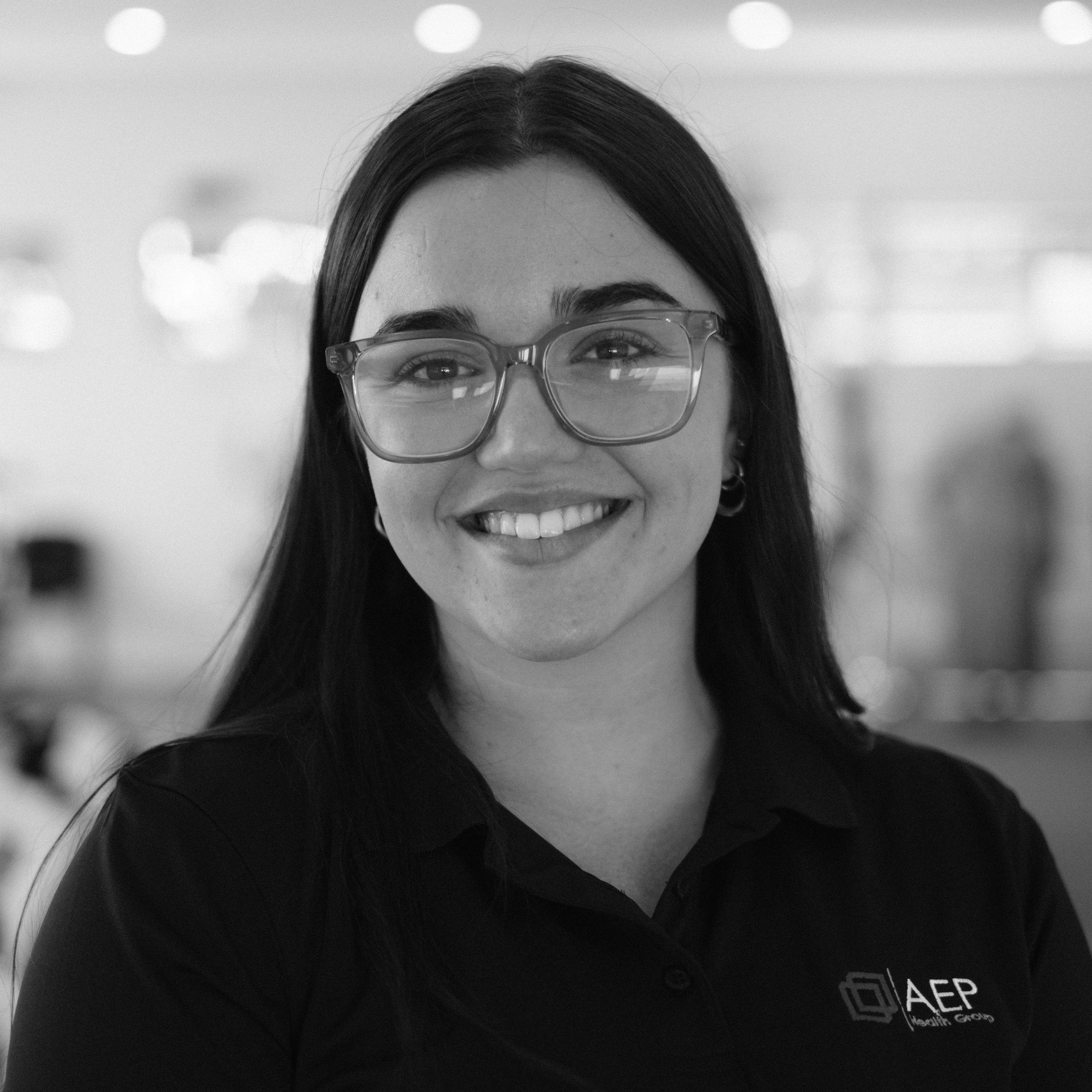 Close-up of a woman with long dark hair, wearing glasses, smiling, with a blurred background.