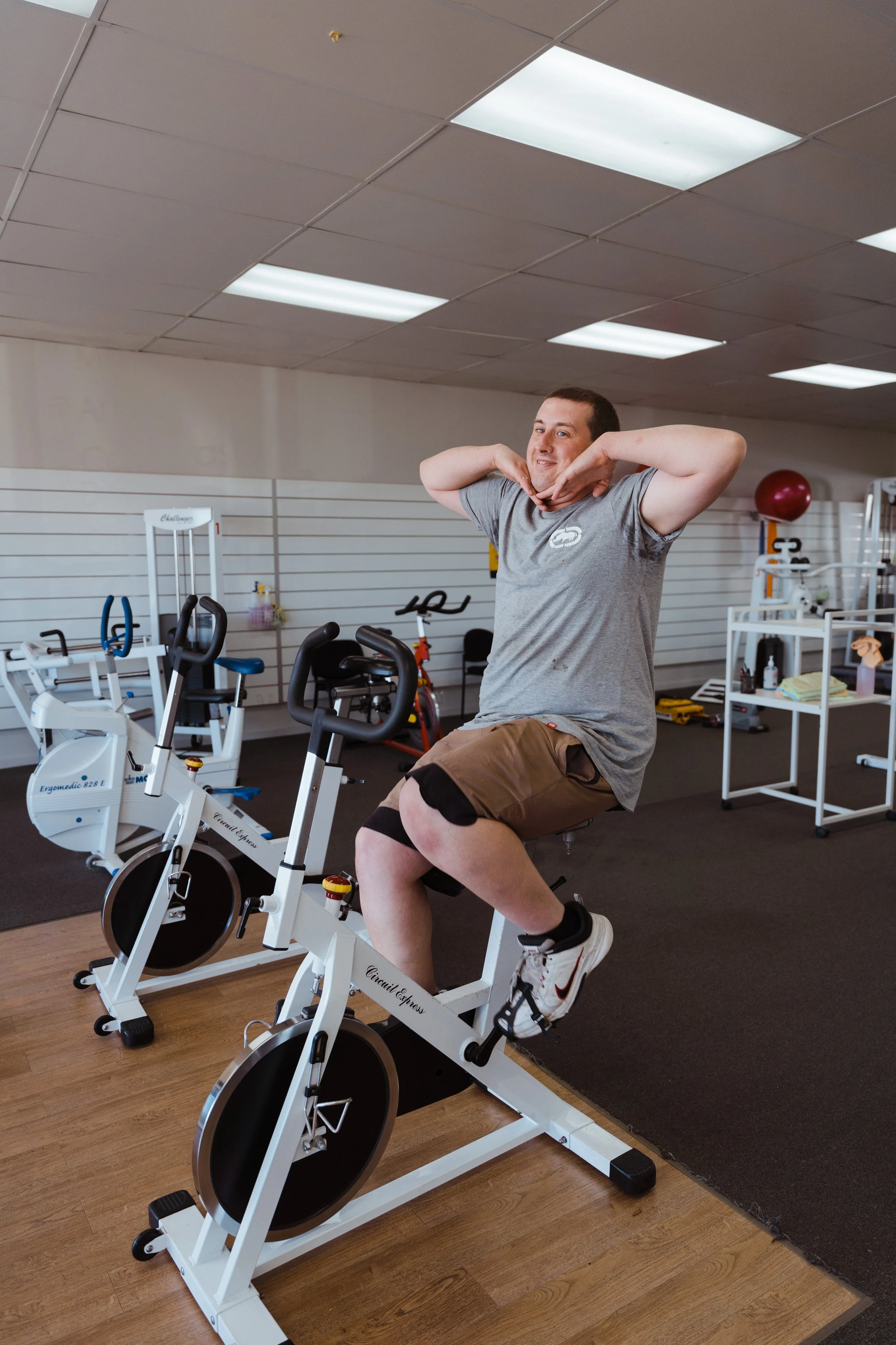 A man smiling and resting his chin on his hands while riding an exercise bike in a fitness room with workout equipment and a white shelf in the background.