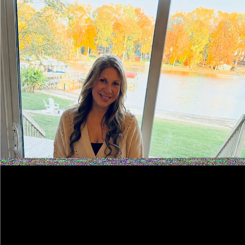 A smiling woman with long, wavy hair standing inside near a large window overlooking a lake and autumn-colored trees.