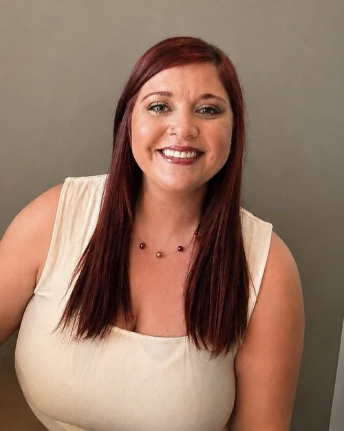 A smiling woman with long red hair wearing a sleeveless cream top and a multicolored beaded necklace posing against a plain gray background.