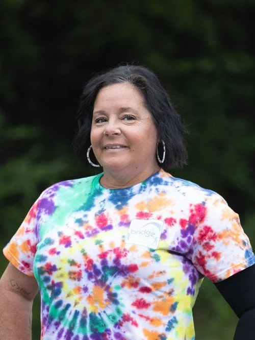 Woman outdoors wearing a colorful tie-dye shirt with a name badge and hoop earrings.