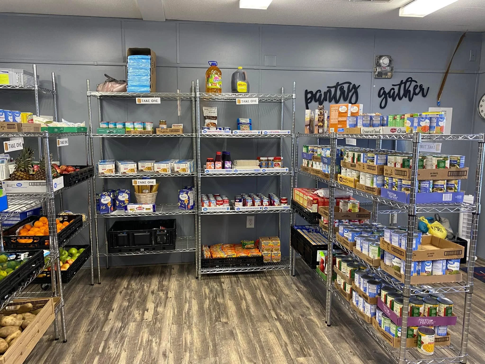 Shelves stocked with canned goods, boxed food, and beverages in a pantry or food bank setting, with signs that say 'Take One' and the words 'pantry' and 'gather' on the wall.