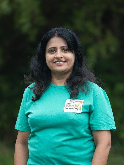 woman with black hair wearing a teal t-shirt with 'the bridge academy' logo standing outdoors in front of green trees