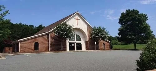 Exterior of a red brick church with a large white arched entryway and a wooden cross on top. Two flowering trees stand in front against a blue sky.