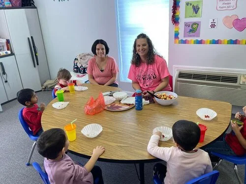Children and two women sitting around a table in a classroom, eating snacks.
