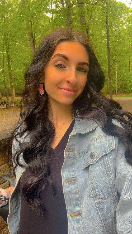 A young woman with long dark wavy hair taking a selfie outdoors in a wooded area. She is wearing a denim jacket, a black top, and colorful earrings, and has a slight smile on her face.