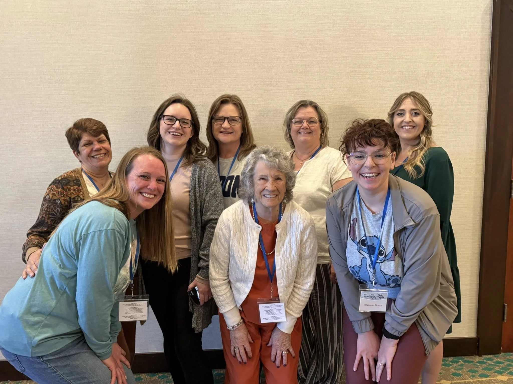 Group of nine women posing together indoors, smiling for a photo at a conference or event, each wearing a name tag.