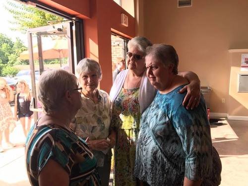 Four elderly women are gathered and talking inside a cafe. One woman is hugging another, and the scene is lit by natural light from the open door.