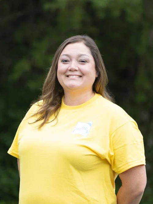 A woman with long brown hair smiling outdoors in front of green trees, wearing a yellow t-shirt.