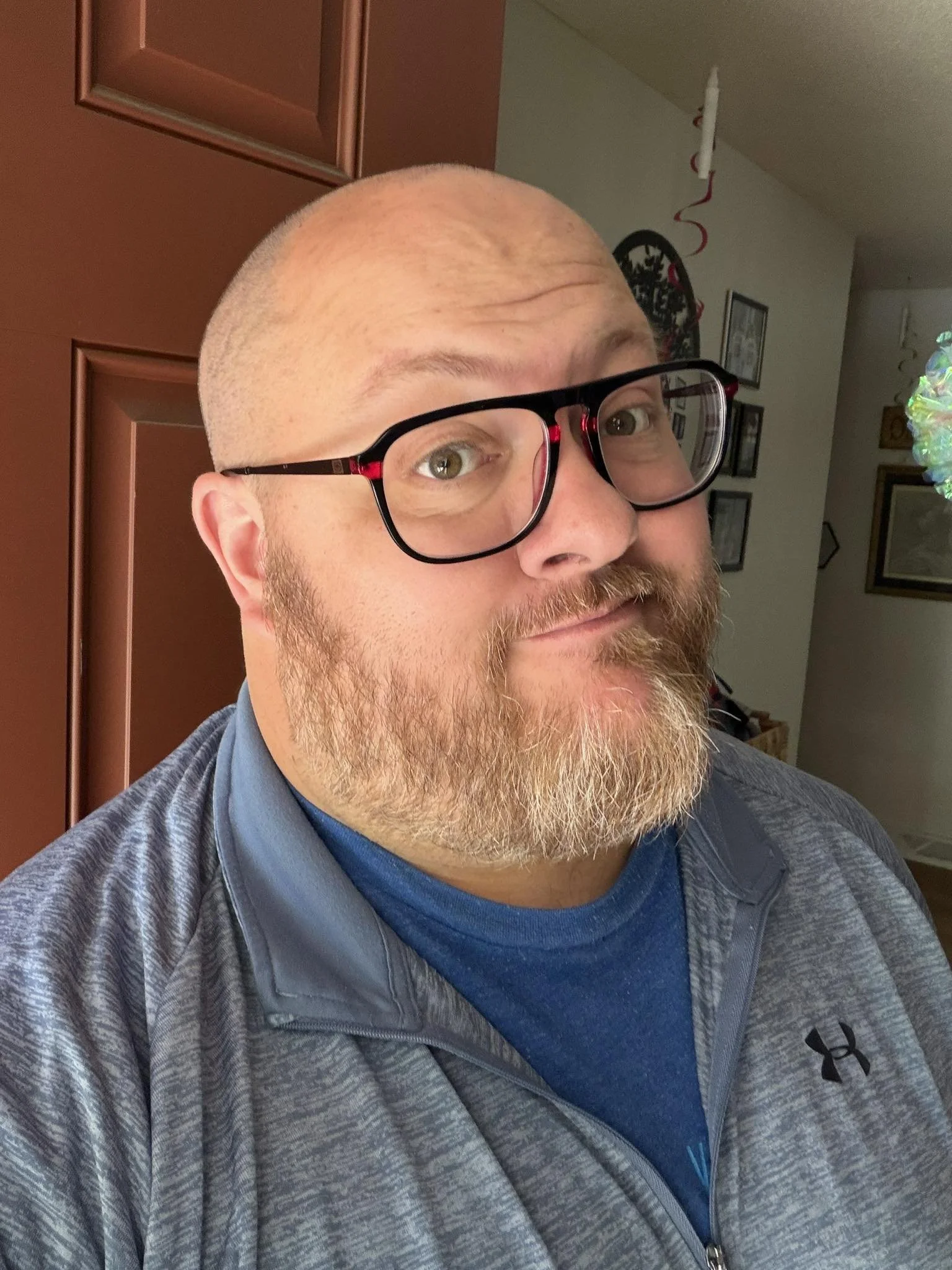 A close-up portrait of a man who is Pastor Brandon; with glasses, a tan beard, and a shaved head, wearing a gray Under Armour jacket over a blue shirt, indoors with framed pictures and decorations on the wall.
