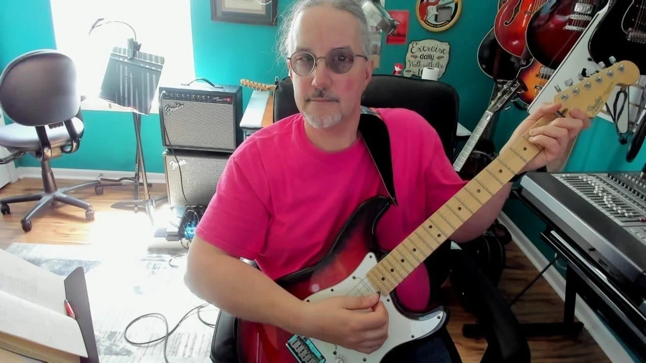A man who is the worship leader Jeff Imbody; with glasses and gray hair playing an electric guitar in a music room, surrounded by musical equipment and guitars on the wall.