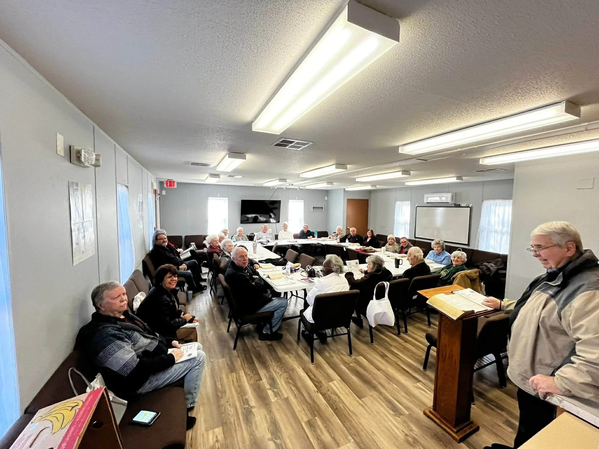 A group of seniors gathered in a meeting room, sitting around a long table, some reading or writing, with a few standing near the speaker's podium.