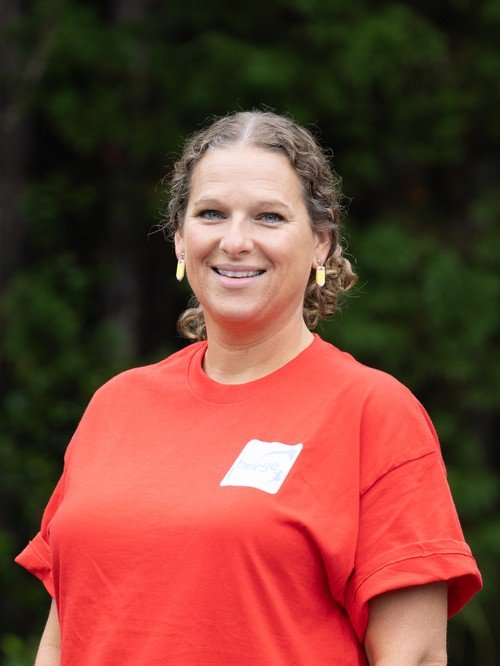 A smiling woman with curly brown hair, wearing a red T-shirt with a name tag and earrings, standing outdoors with green trees in the background.