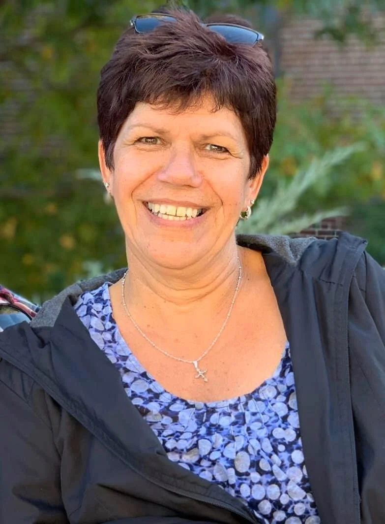 A smiling woman with short dark hair, wearing sunglasses on her head, a necklace with a starfish pendant, earrings, a patterned blue shirt, and a black jacket, standing outdoors with trees and greenery in the background.