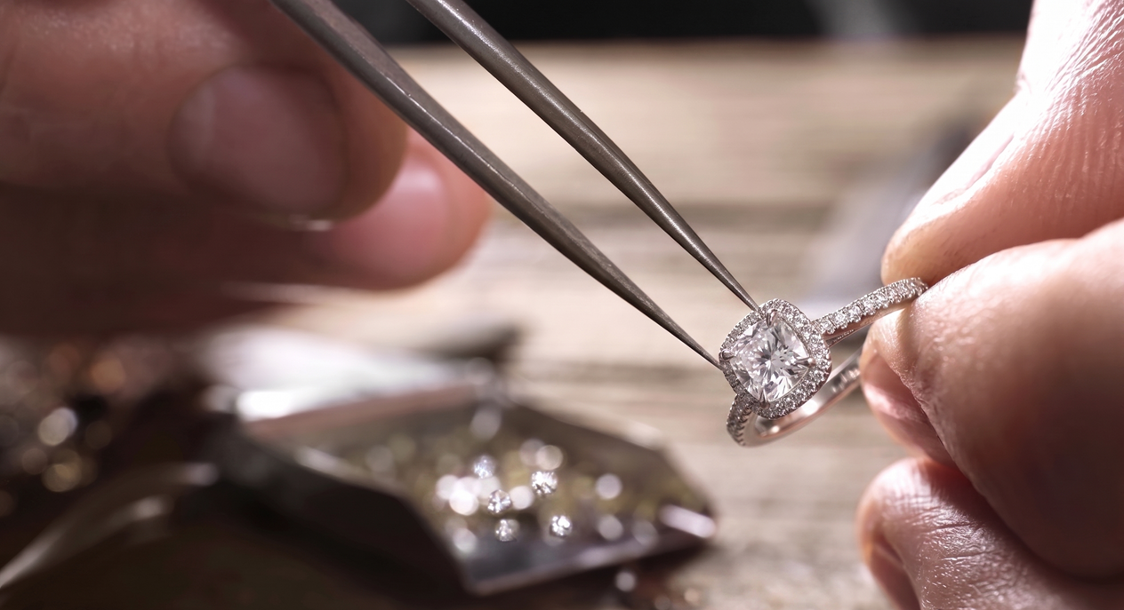 A person using tweezers to handle a diamond engagement ring with a large central diamond and small surrounding diamonds.