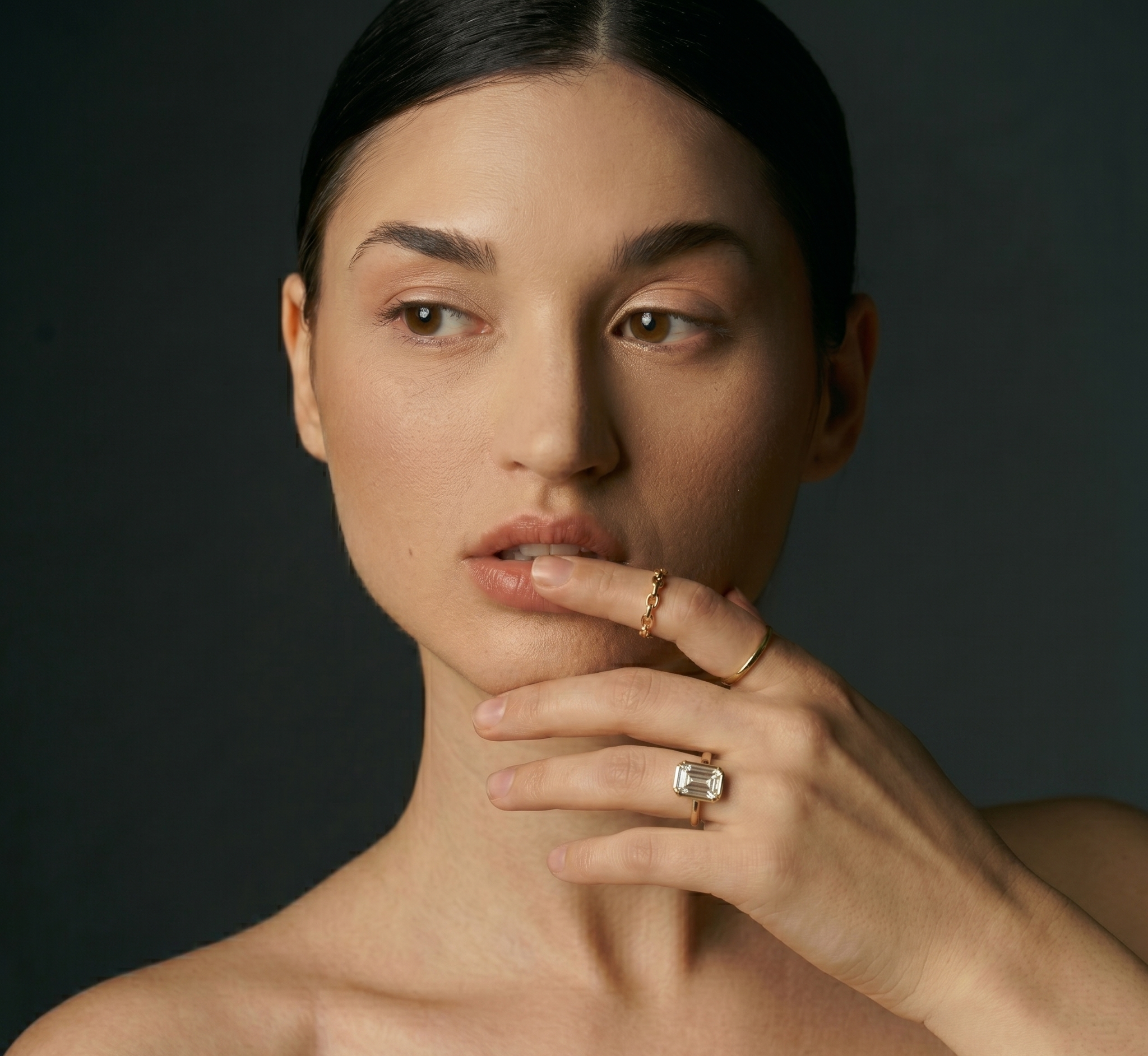 Close-up of a woman with dark hair and light skin, touching her face with her hand, wearing jewelry including a large ring, a thinner band, and two rings on her fingers, set against a dark background.