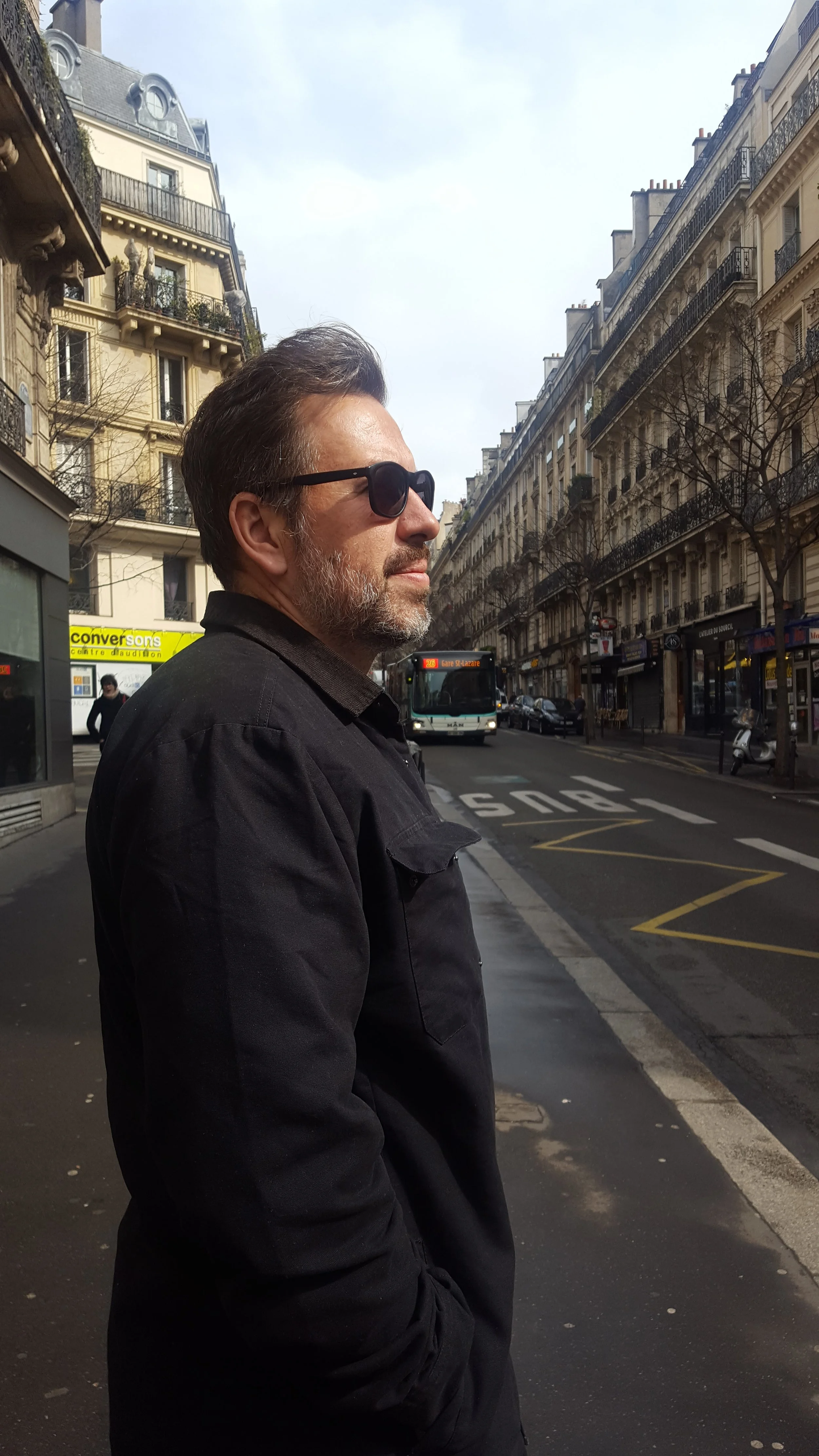 A man wearing sunglasses and a black jacket standing on a city street with tall buildings, a bus, and a few pedestrians in the background.