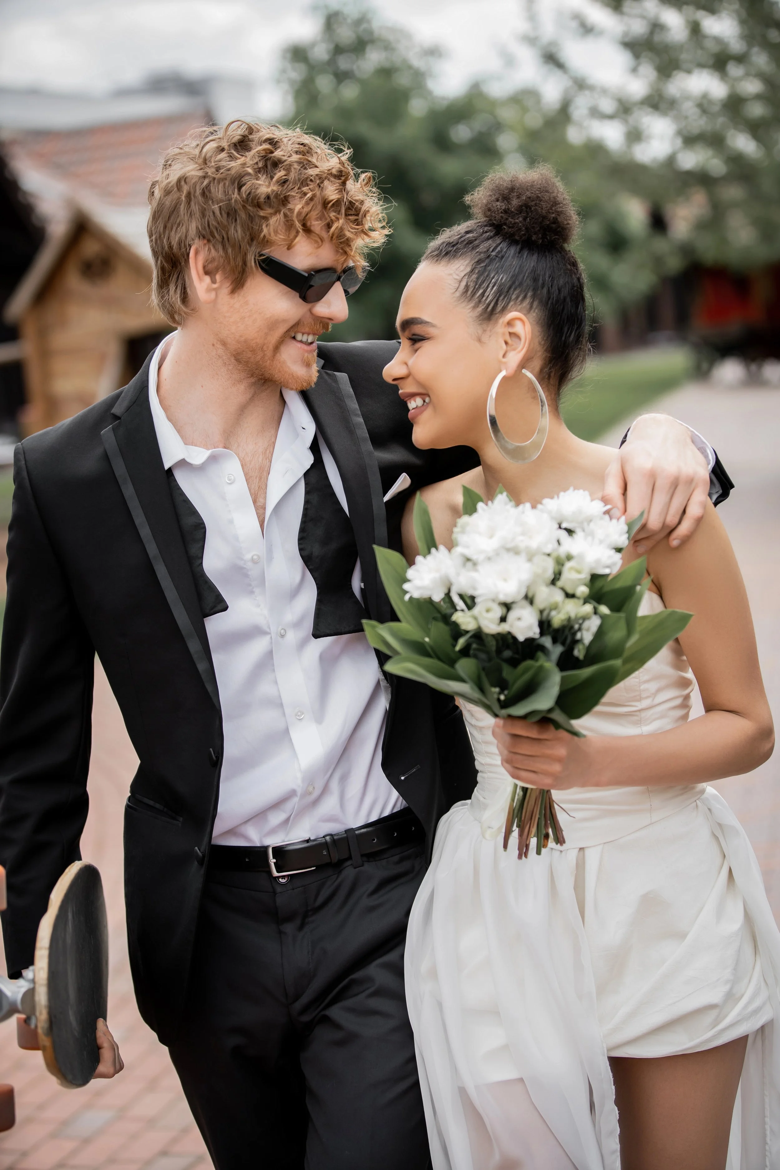 A man and a woman smiling and looking at each other, with the woman holding a bouquet of white flowers. The man is wearing sunglasses and a black suit with a white shirt. The woman has large hoop earrings and is wearing a light-colored dress with a high slit.