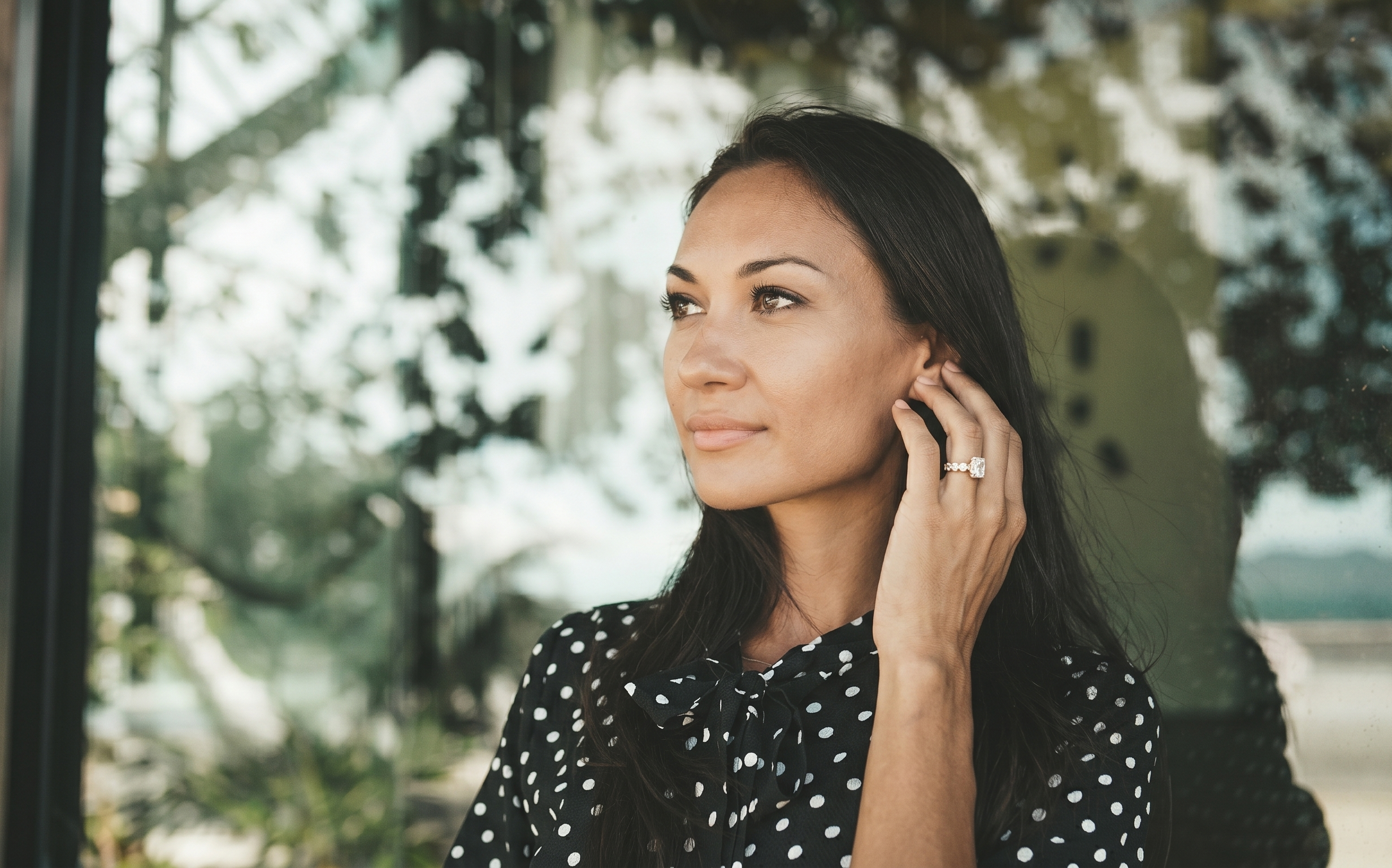 A woman with long dark hair, wearing a black polka dot top, is touching her ear and looking slightly to the right, outdoors with greenery reflected in the window behind her.