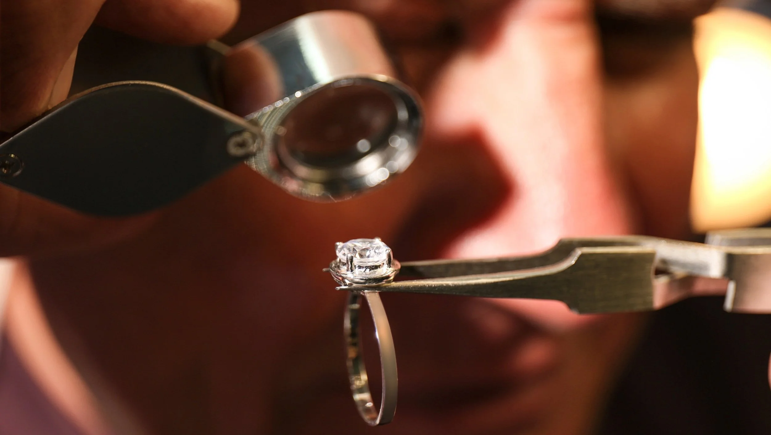 A jeweler's hand holding a magnifying glass inspecting a diamond ring during a setting process.